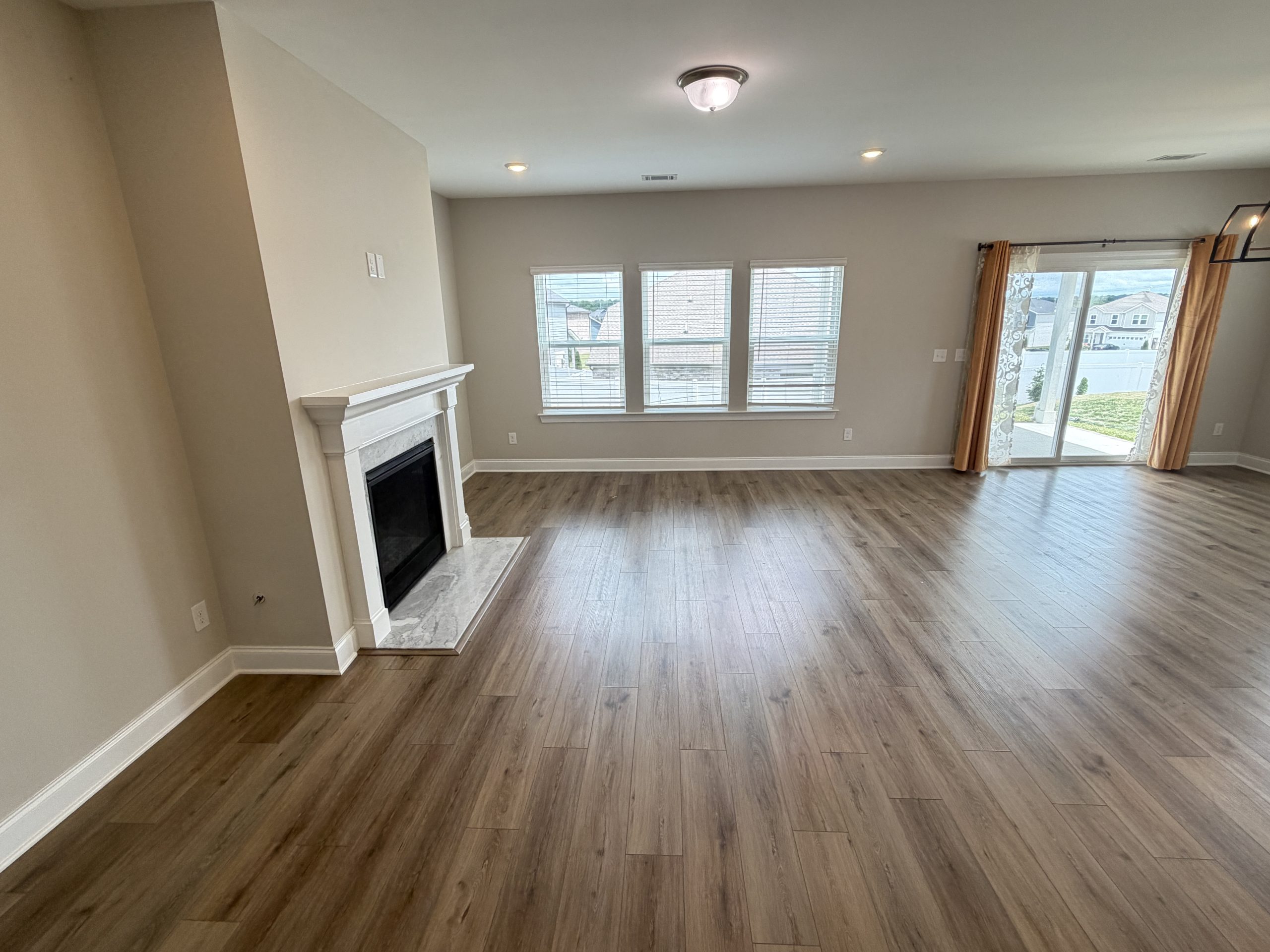 Spacious living room with a white fireplace, light wood flooring, three front-facing windows, and a sliding glass door with mustard curtains to an outdoor view.