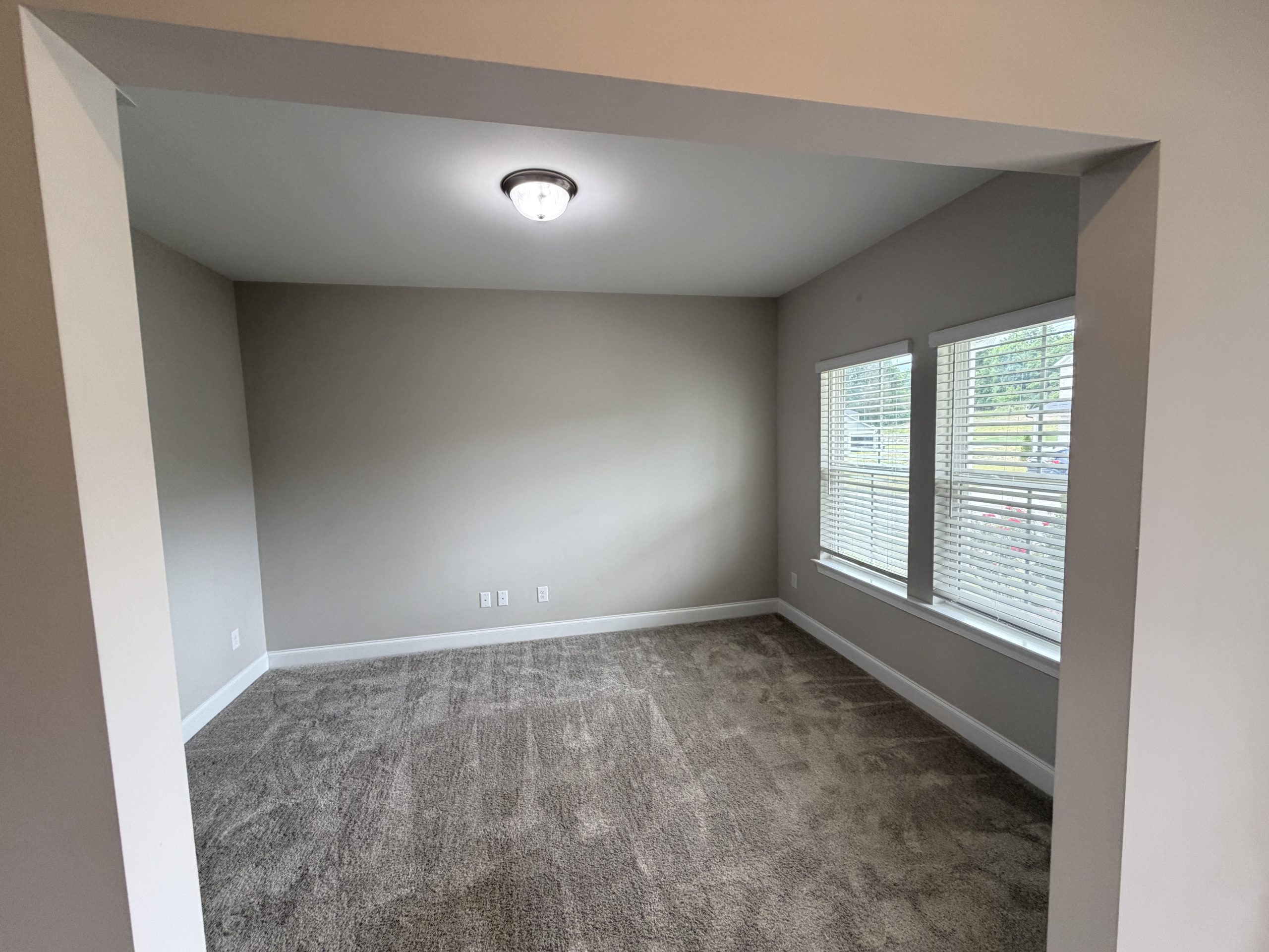 Empty room with gray walls, beige carpet, a central ceiling light, and a large window with blinds casting natural light into the space.