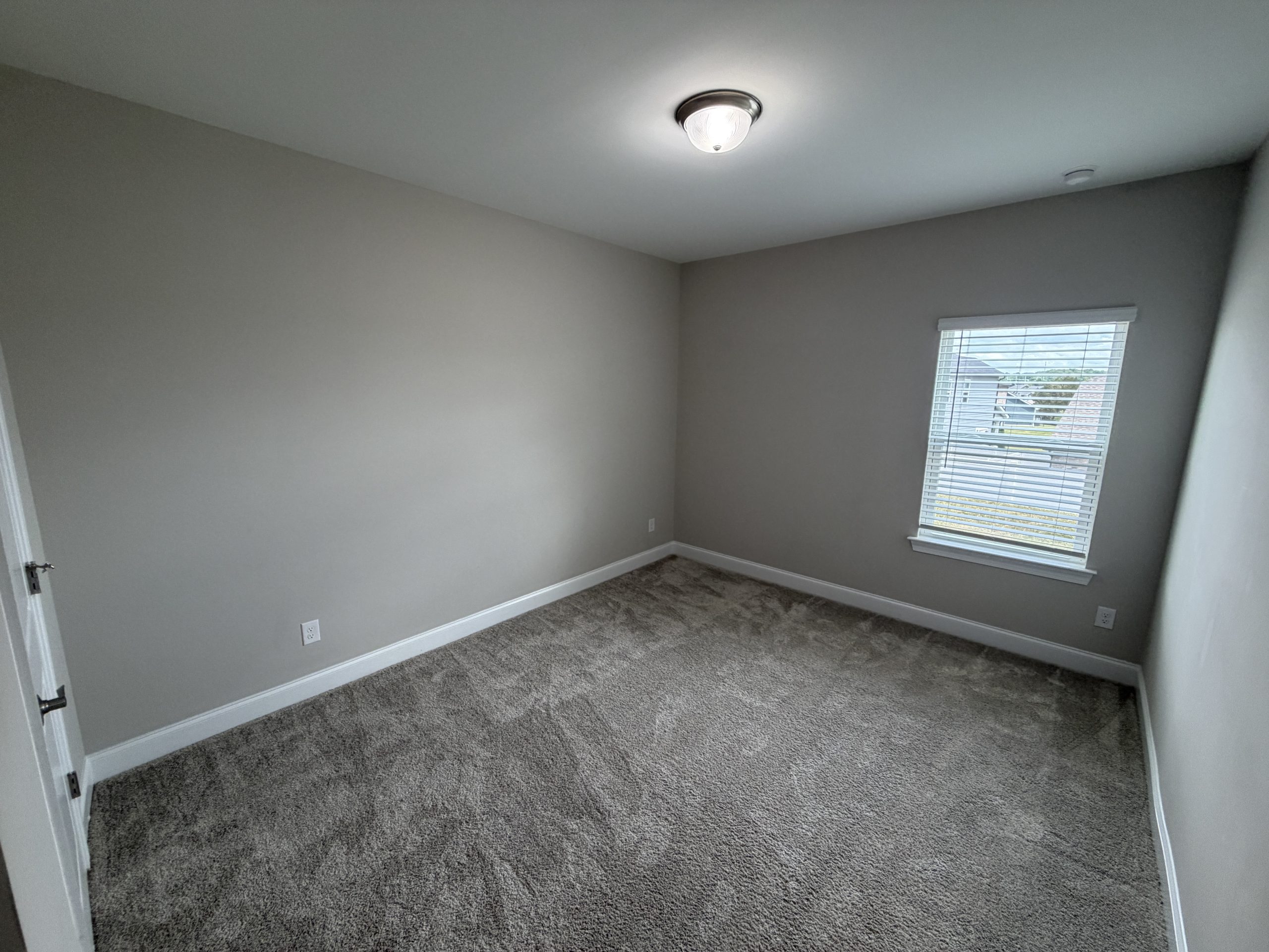 Empty small bedroom with beige walls, gray carpet, a window with blinds, and a ceiling light fixture.