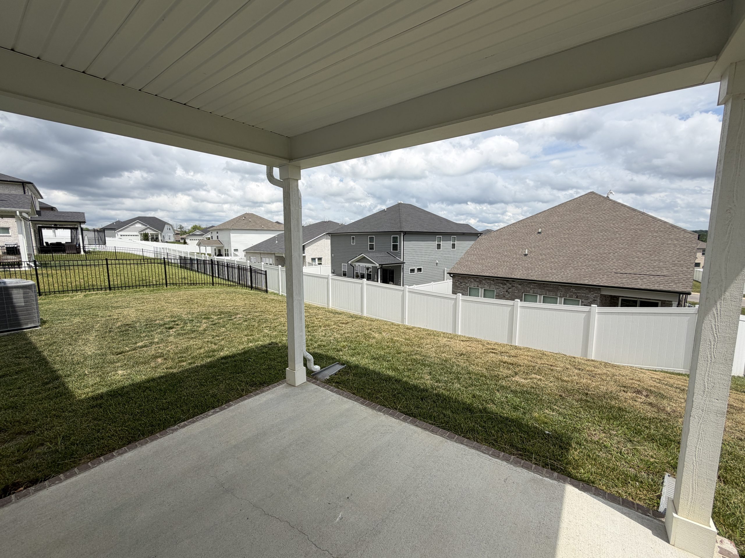 Backyard view from under a covered patio: white posts, green grass, and a white fence with neighboring houses beyond under a cloudy sky.