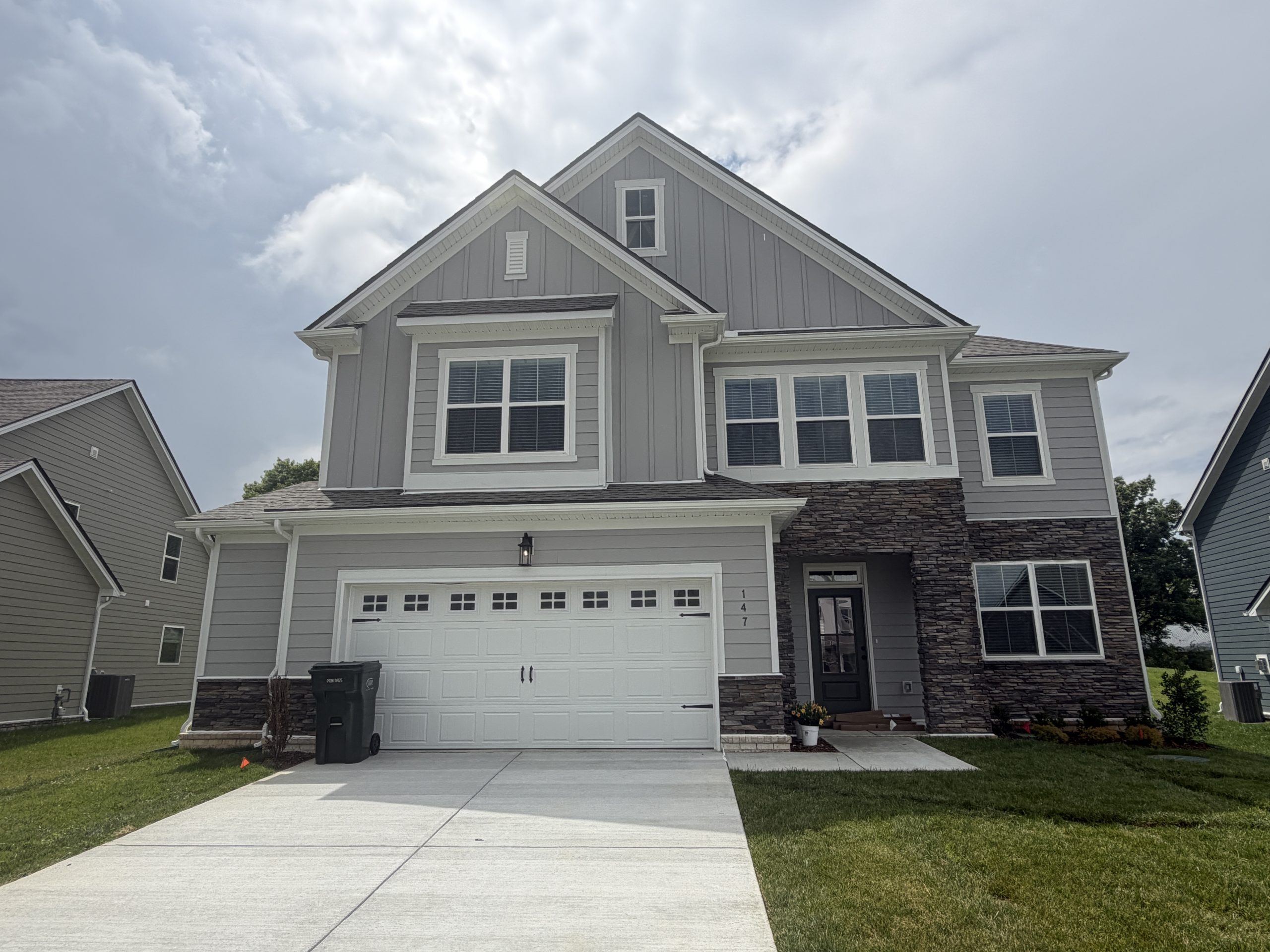 Two-story gray house with white trim, stone accents, and a white garage door seen from the front yard and driveway.