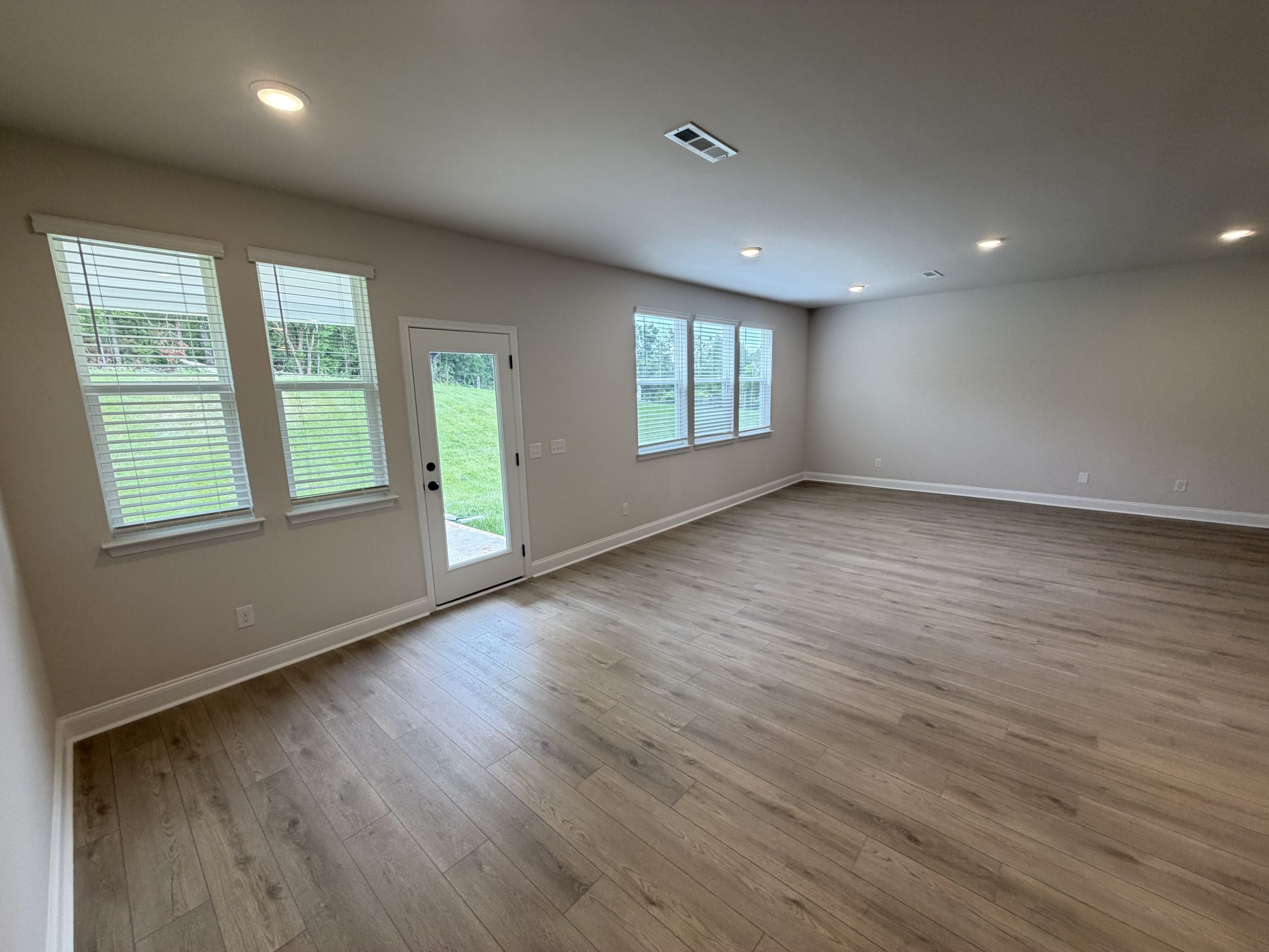Empty living room with light wood flooring, gray walls, multiple windows with blinds, and a glass door to the yard.