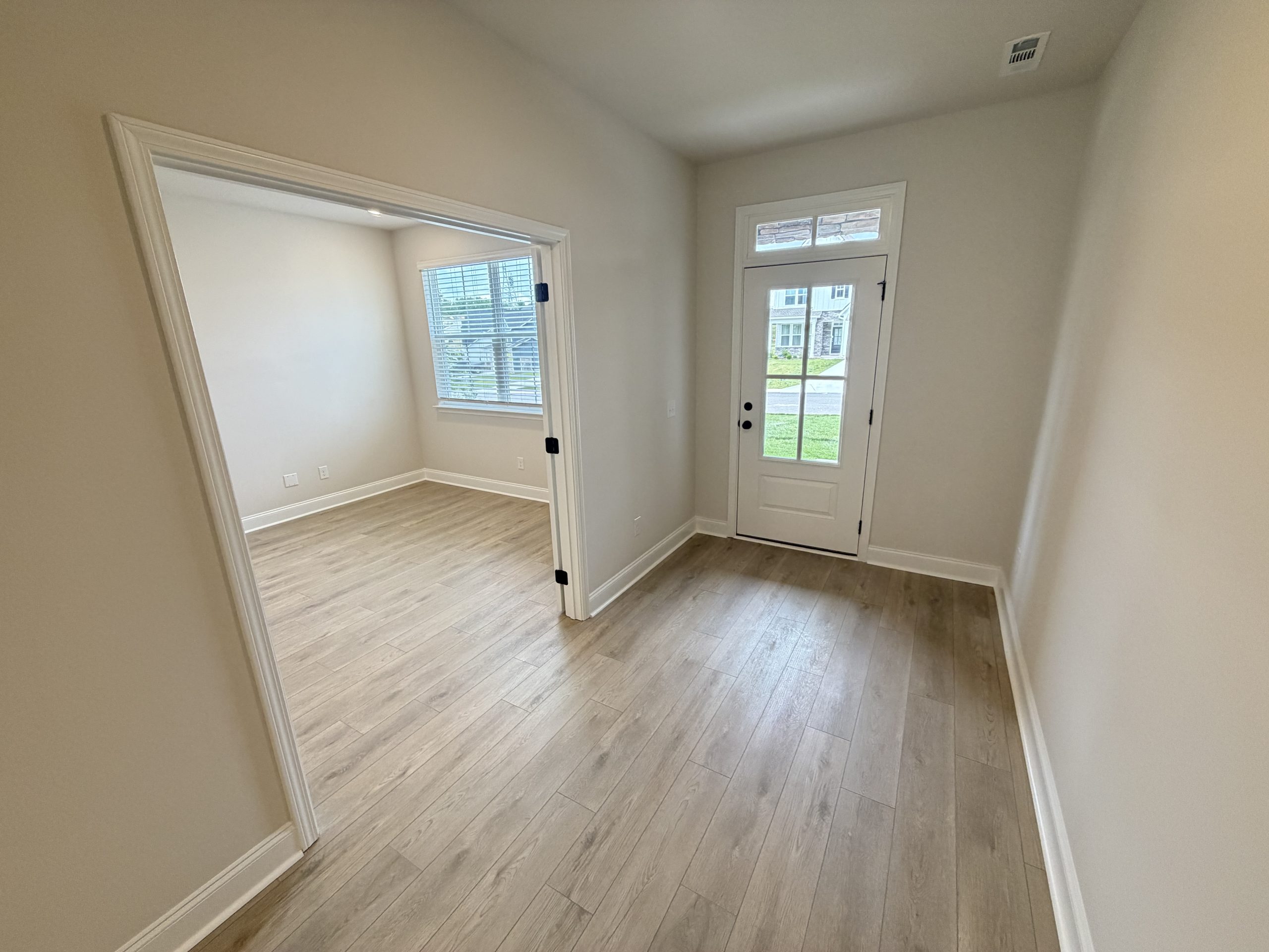 Interior hallway with light wood flooring, beige walls, and a white front door with glass panes visible to the outside. An adjacent room with a window and blinds is seen through an open doorway.