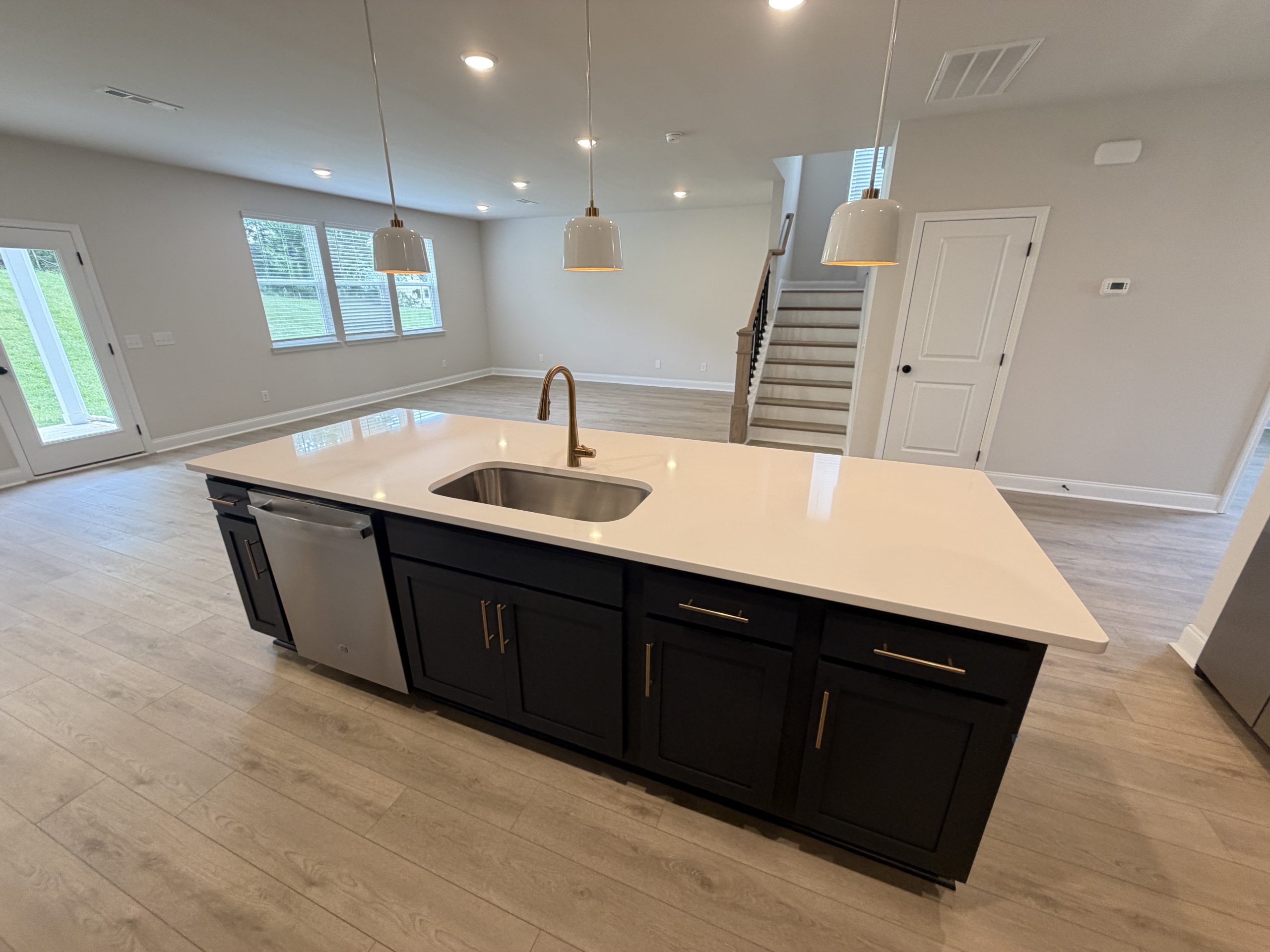 Bright open-concept kitchen with a large white island, brass faucet, and dark lower cabinets; light gray walls and wide plank flooring visible, plus a staircase in the background.