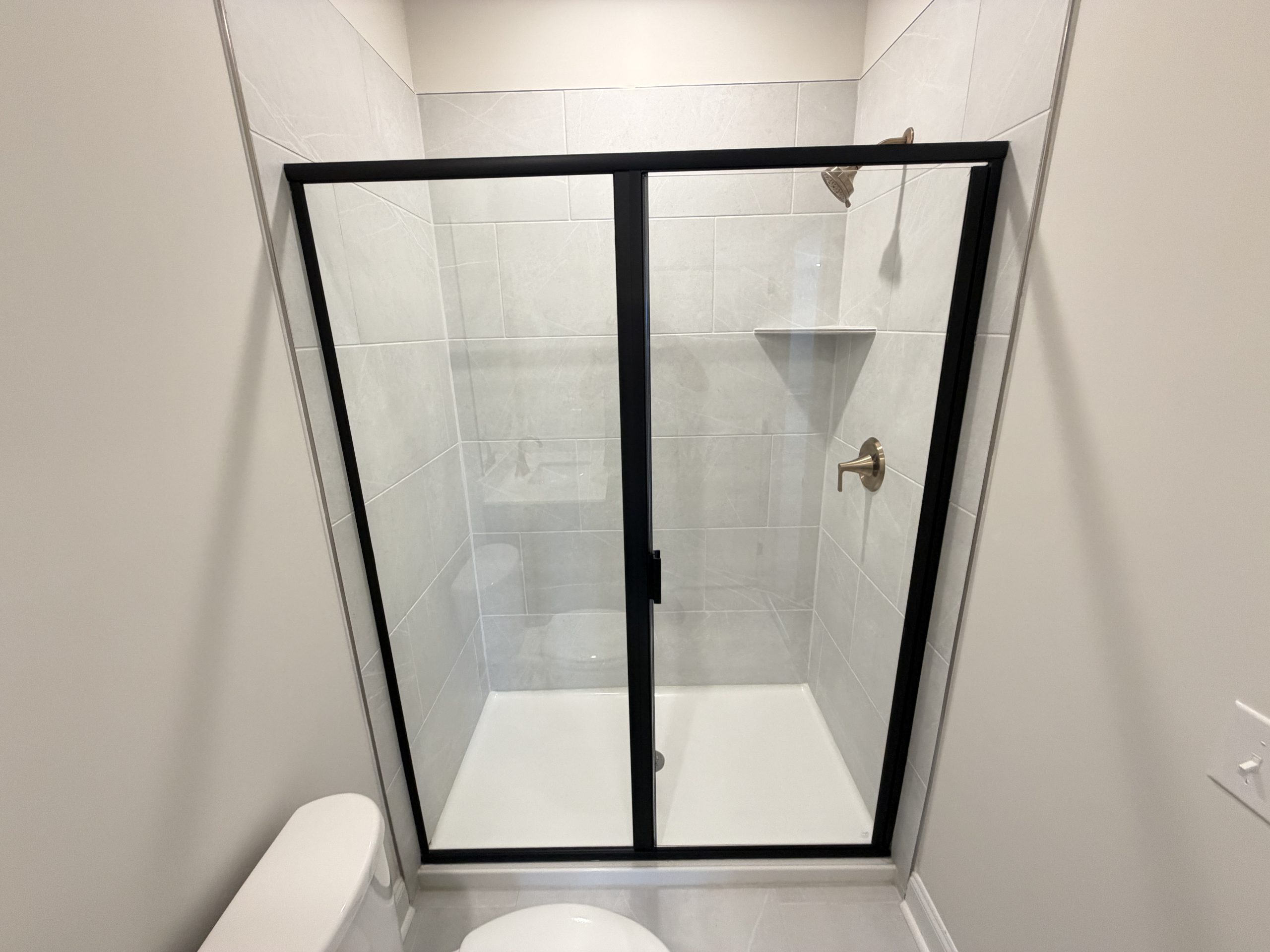Tall view of a small bathroom with a sliding glass shower door framed in black, gray tiled shower walls, and a toilet visible at the bottom left outside the enclosure.