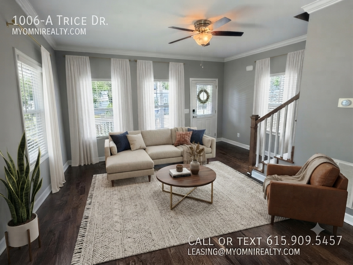 Bright living room in a modern home: gray walls, white curtains, beige sectional sofa, round coffee table, and a staircase with dark wood railing.