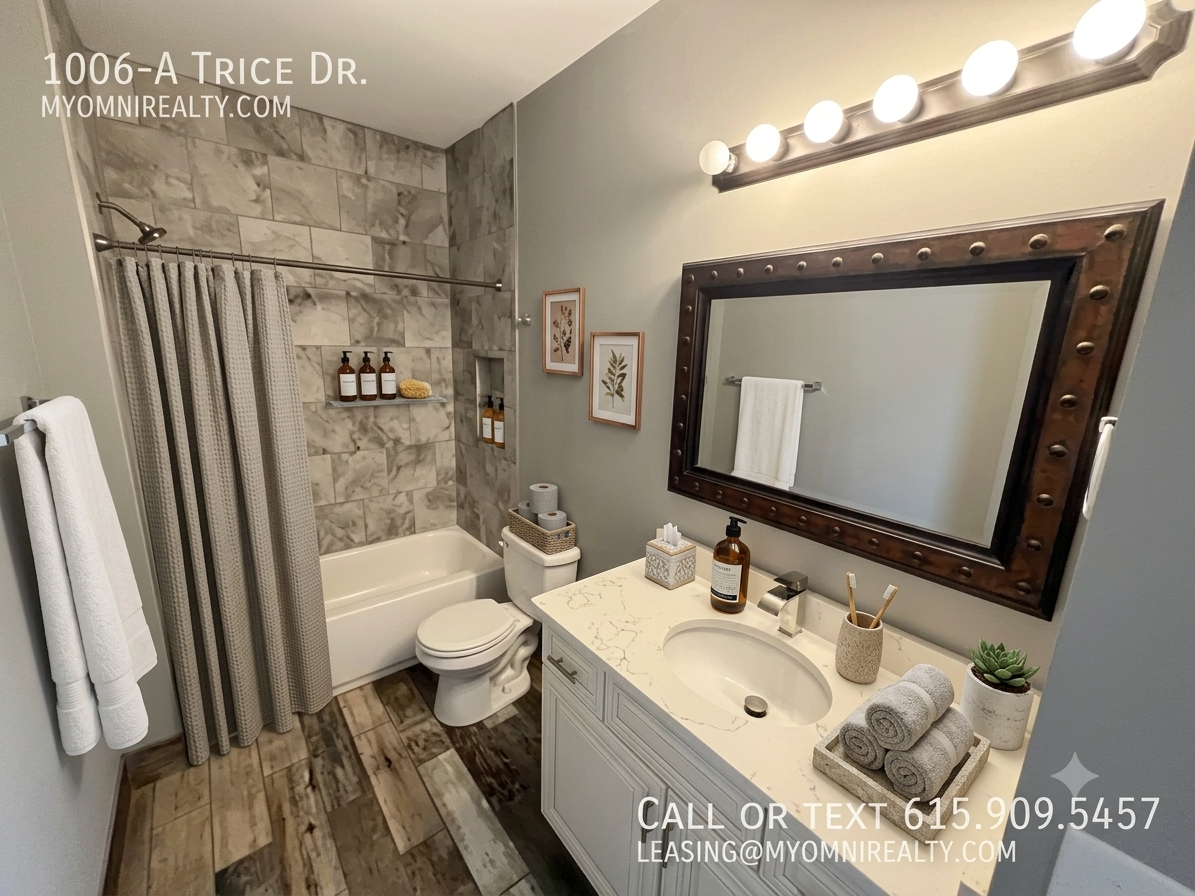 Modern bathroom with marble-tiled shower and tub, gray walls, white vanity with a marble countertop, a large riveted dark-frame mirror, and bright Hollywood-style vanity lights. Includes towels, plants, and brushed bronze accents. The overlay shows a listing address (1006-A Trice Dr.) and contact info.