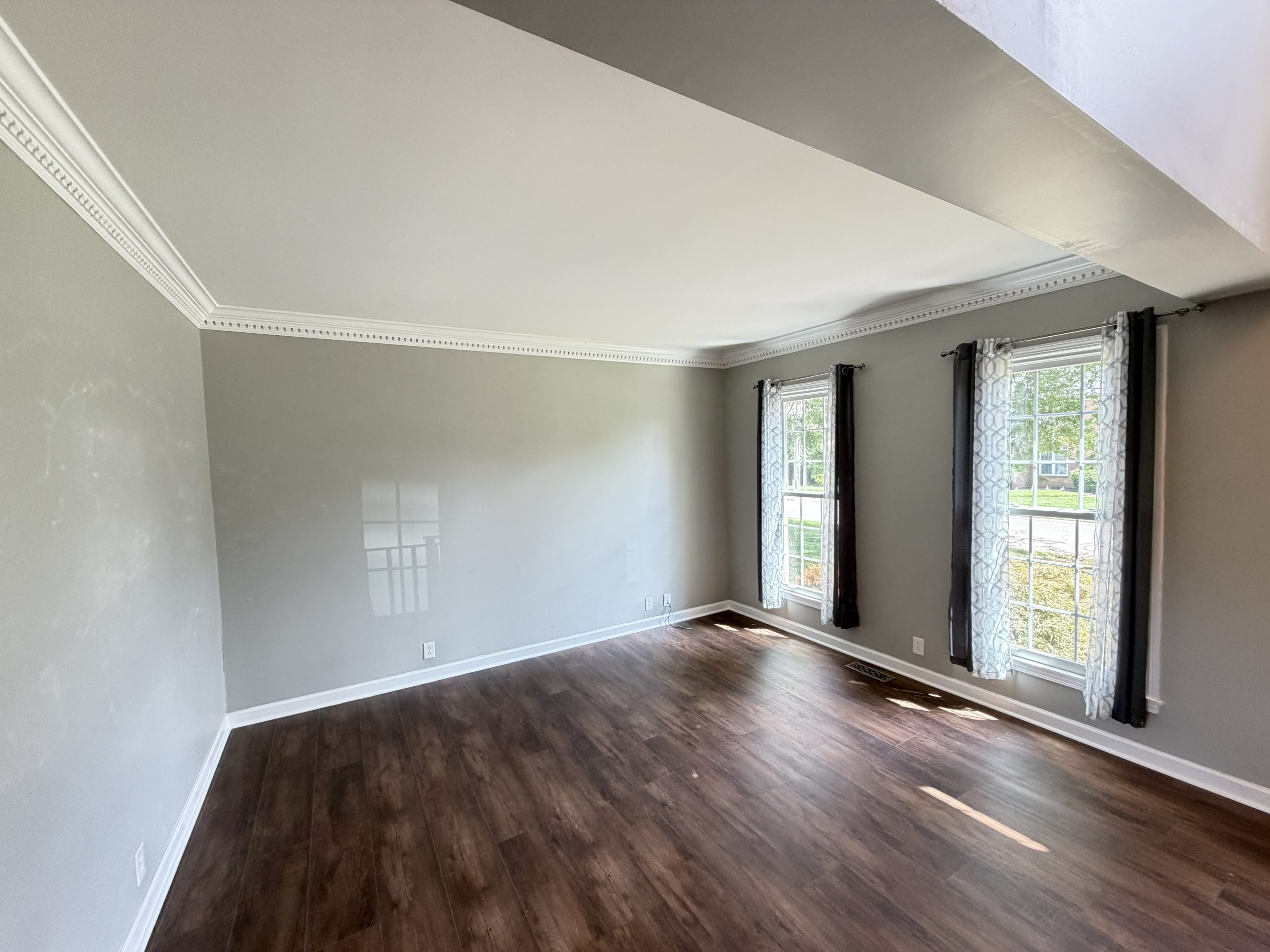 Empty living room with gray walls, dark wood floor, and large windows with patterned curtains.