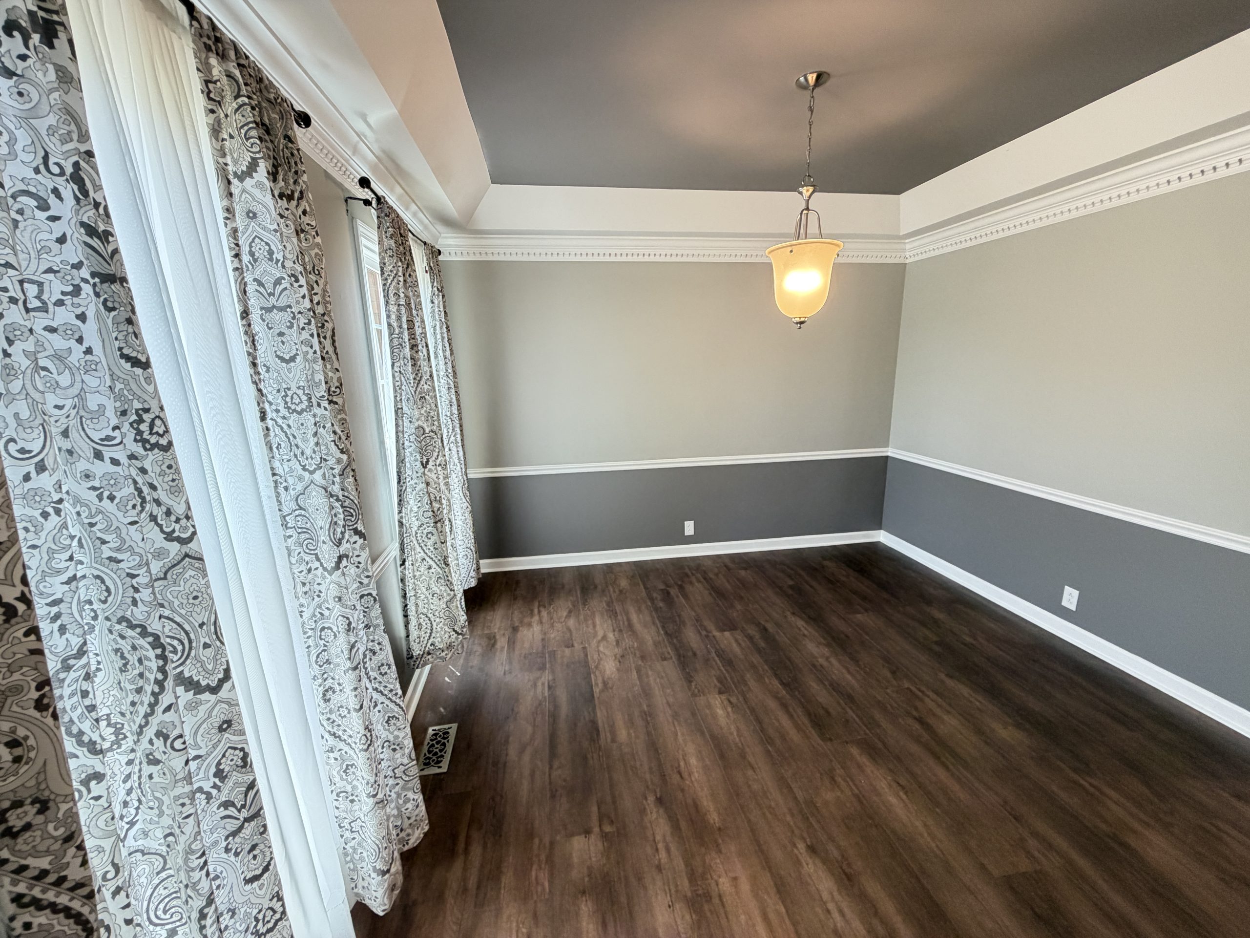 Corner of a room with gray walls, white crown molding, and a hanging pendant light; patterned curtains along left side and dark wood floor.