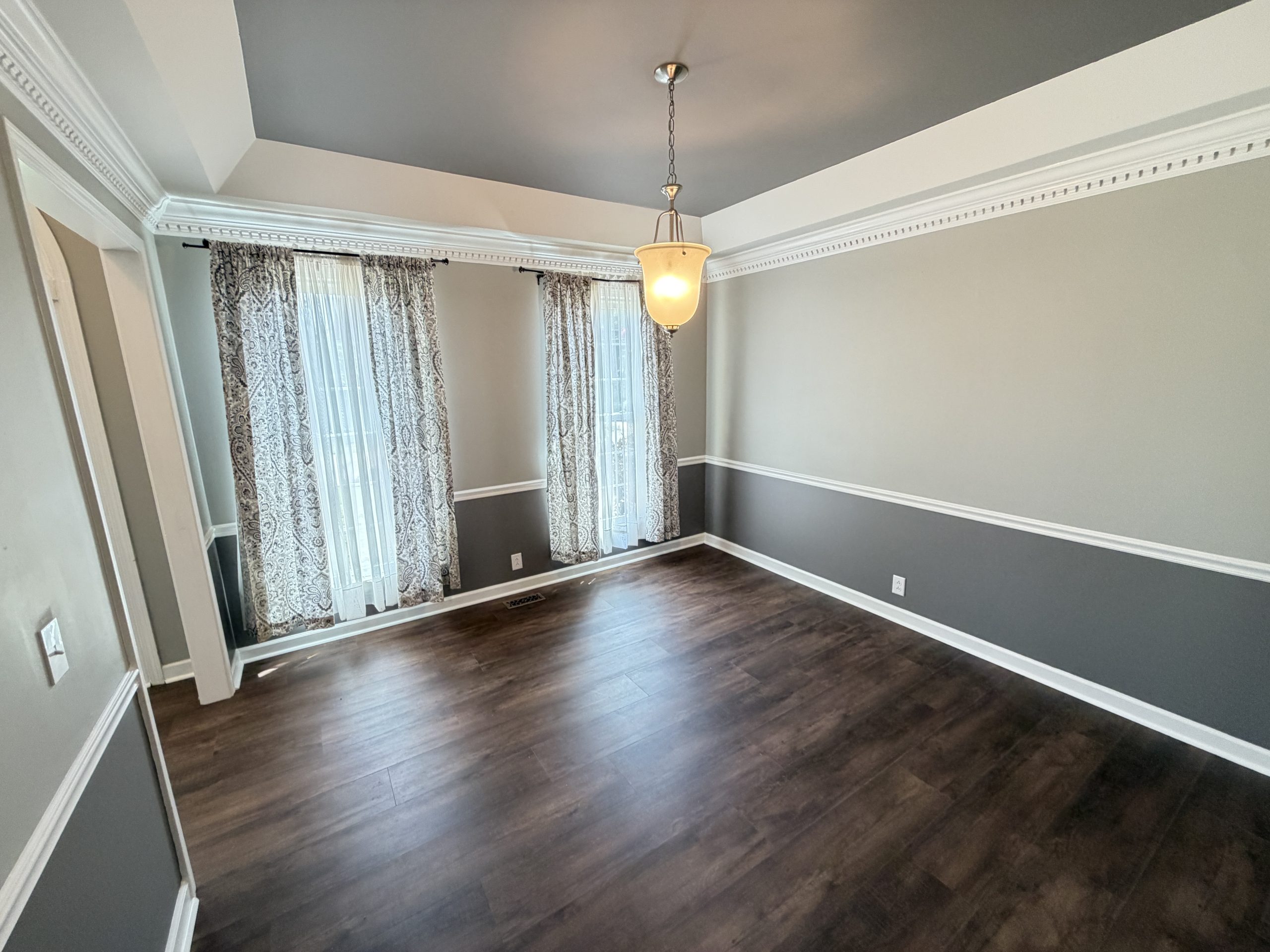 Empty dining room with gray walls, white crown molding, dark wood floor, hanging pendant light, and patterned curtains over tall windows.