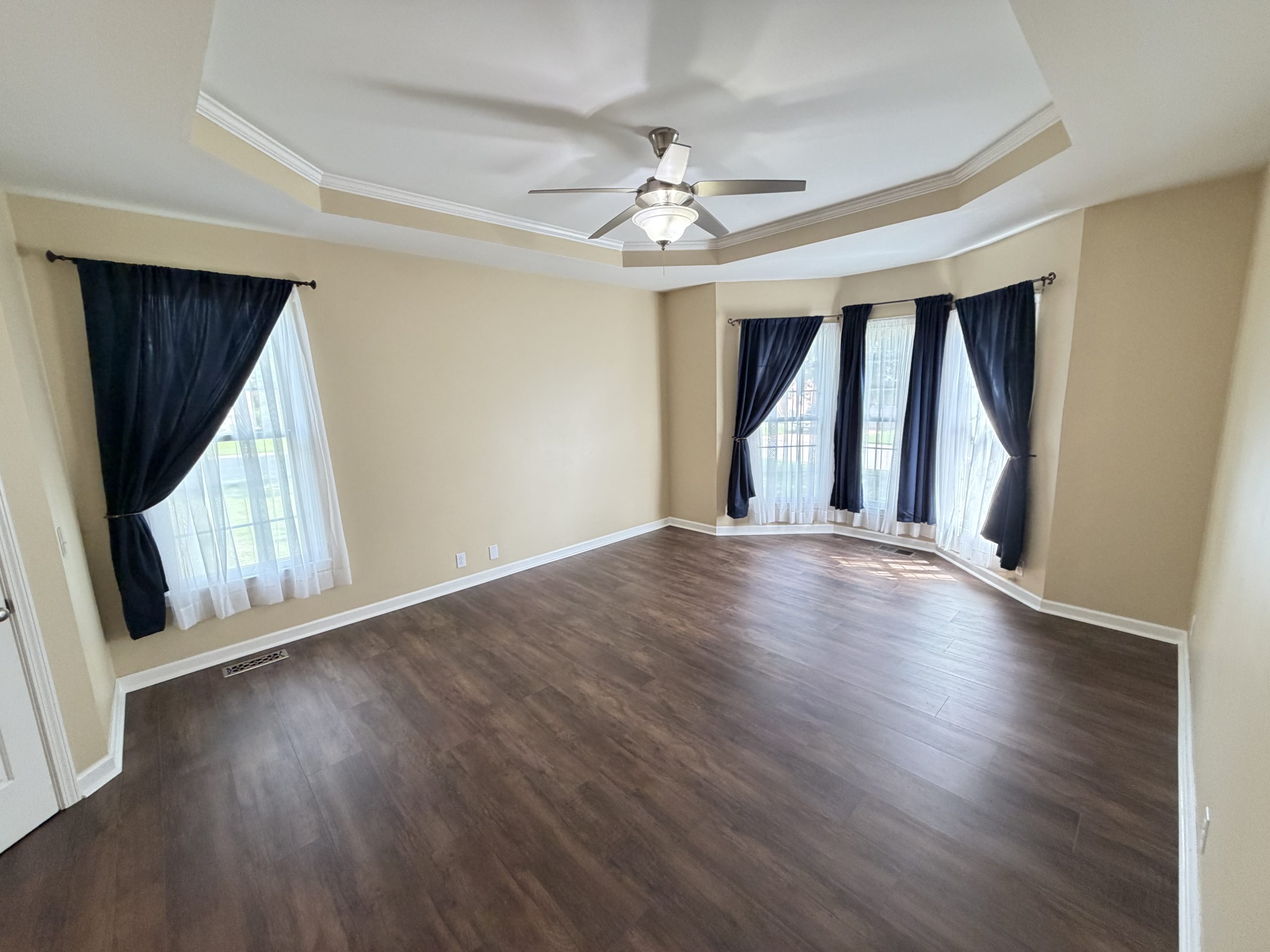 Empty living room with beige walls, dark wood floors, and a tray ceiling featuring a ceiling fan; windows with navy drapes and sheer white curtains.