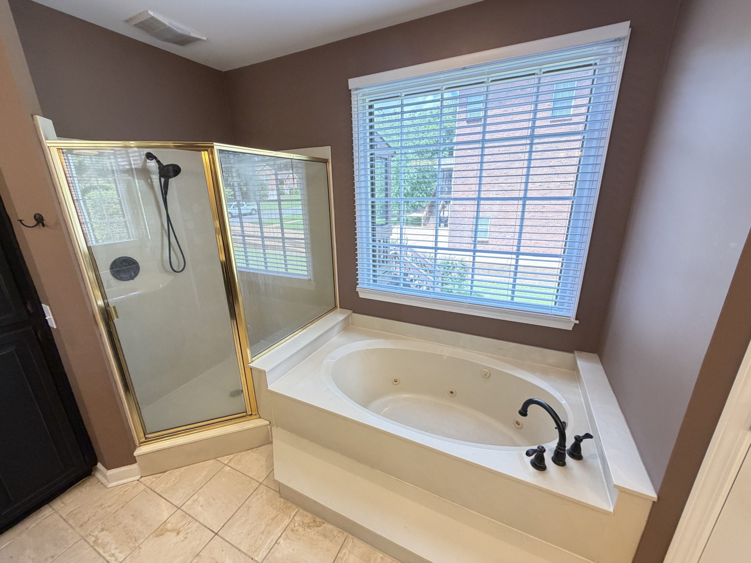 Bathroom with a white soaking tub beside a glass-enclosed shower (gold trim) and a large window with blinds.