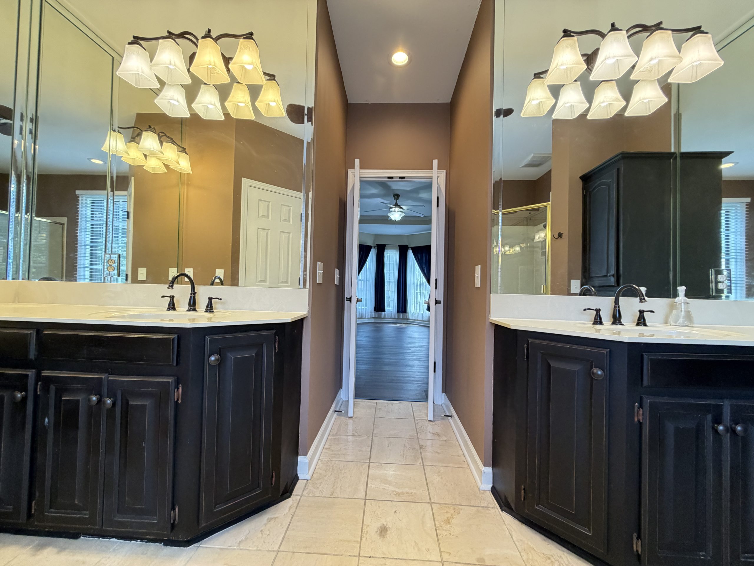Double-sink bathroom vanity with dark cabinets and beige countertop, mirrored walls, and glass-front mirrors reflecting chandelier-style lights behind.