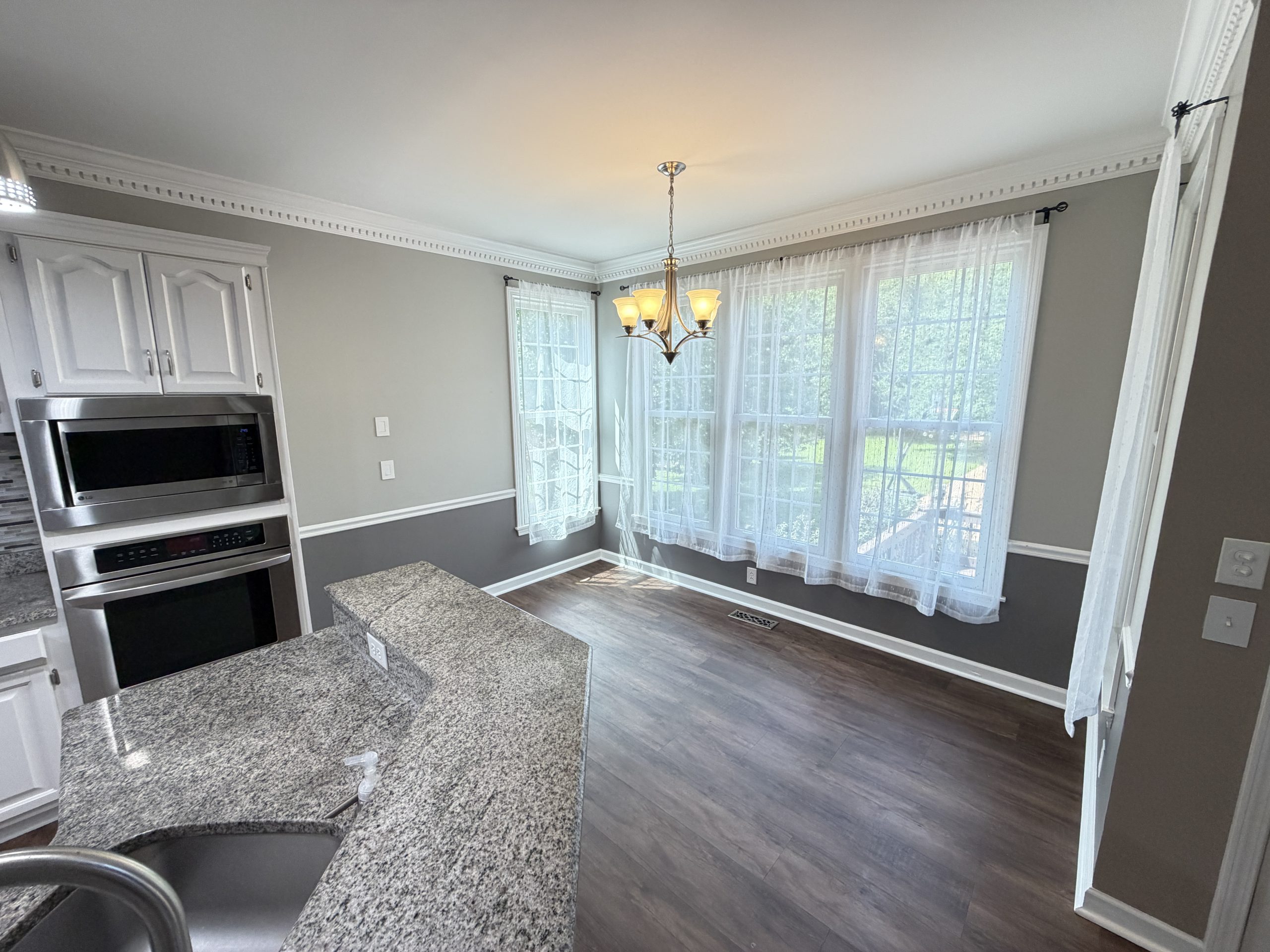 Modern kitchen with granite island, stainless steel double oven, white cabinets, and a chandelier over the dining area by large windows with sheer curtains.