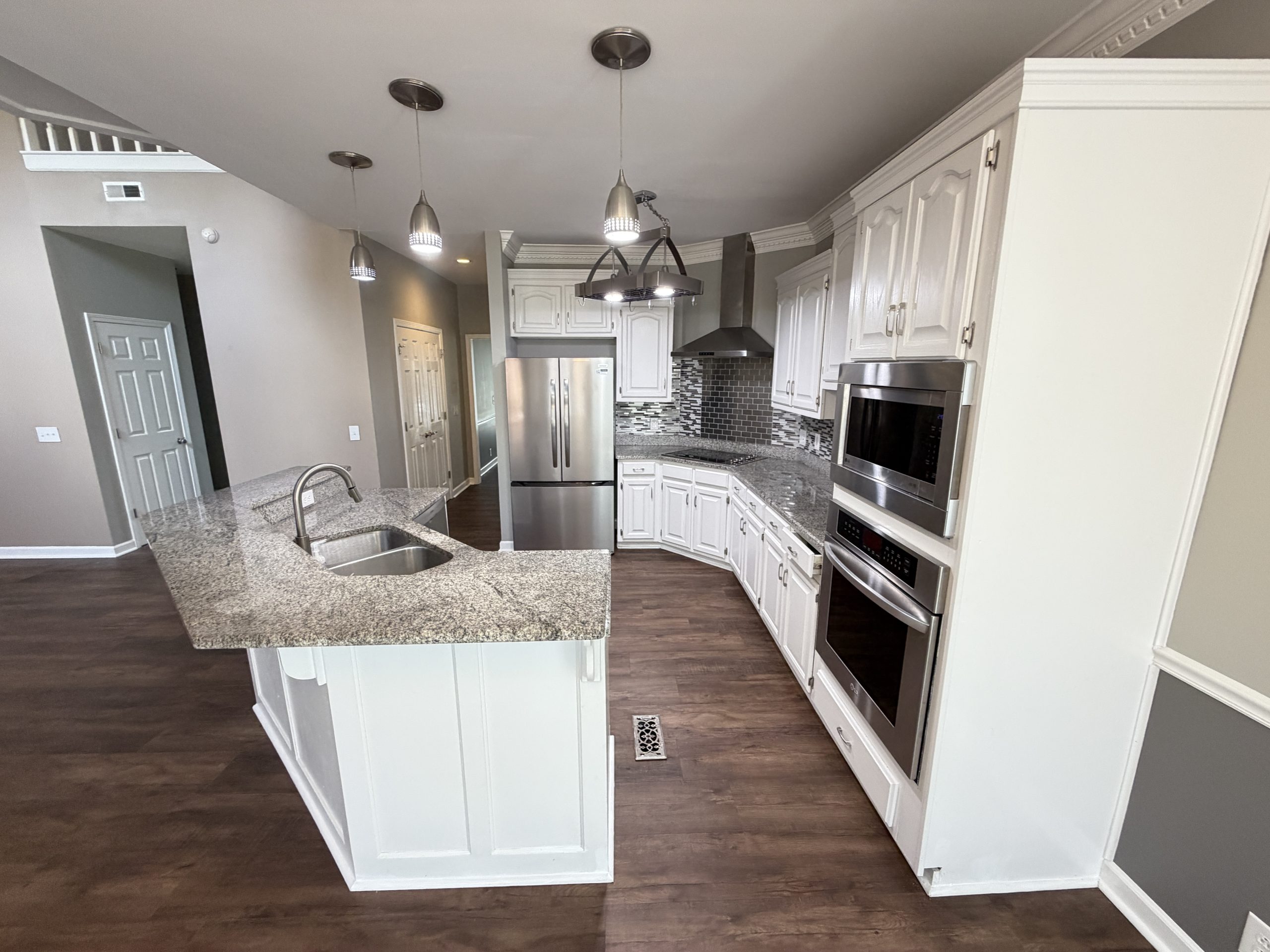 Wide-angle view of a bright white kitchen with granite countertops, a center island, stainless steel refrigerator, and pendant lights.",