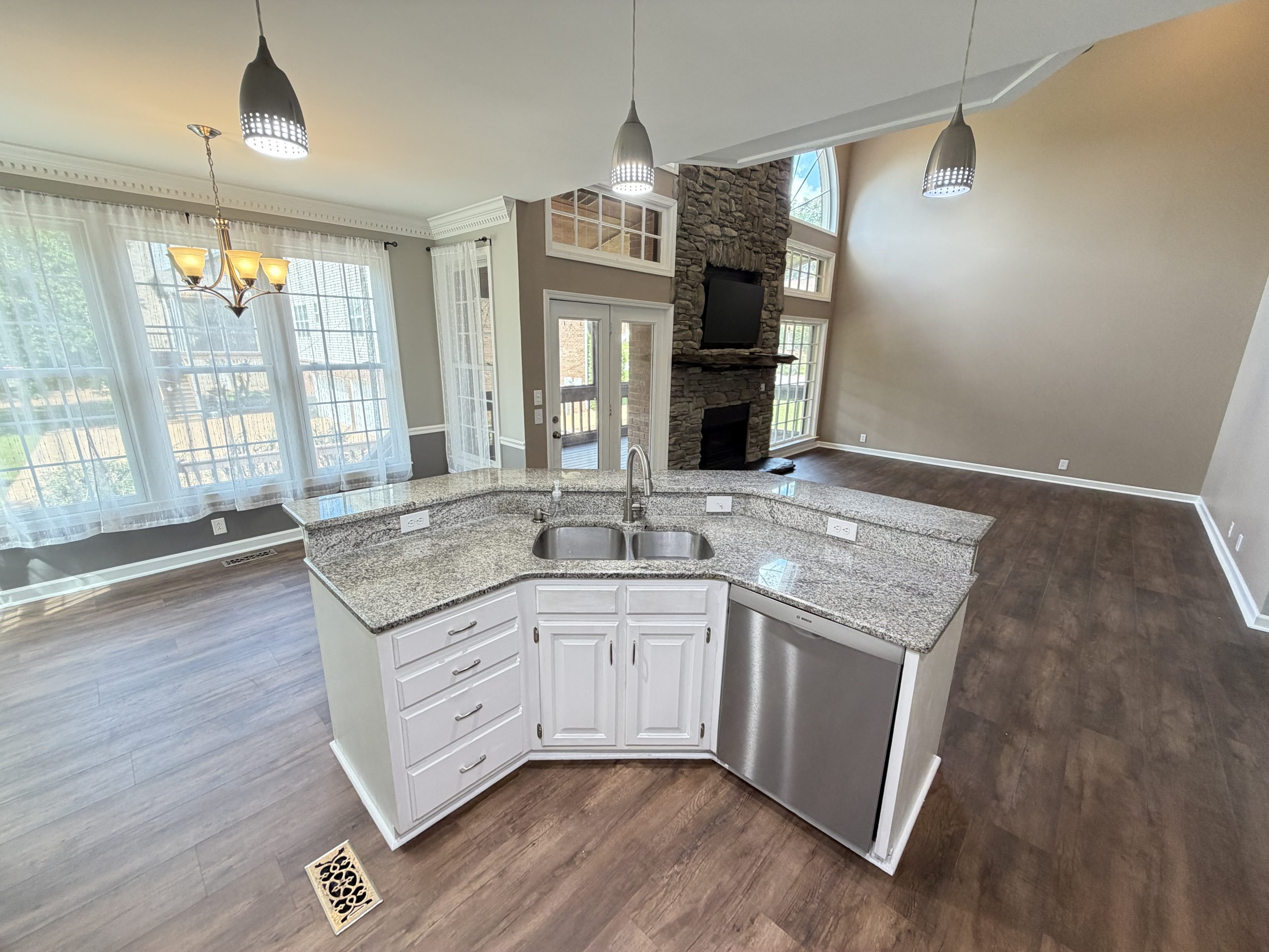 Open-concept kitchen with a granite island, white cabinetry, and a stainless dishwasher, overlooking a living area with a stone fireplace and large windows.