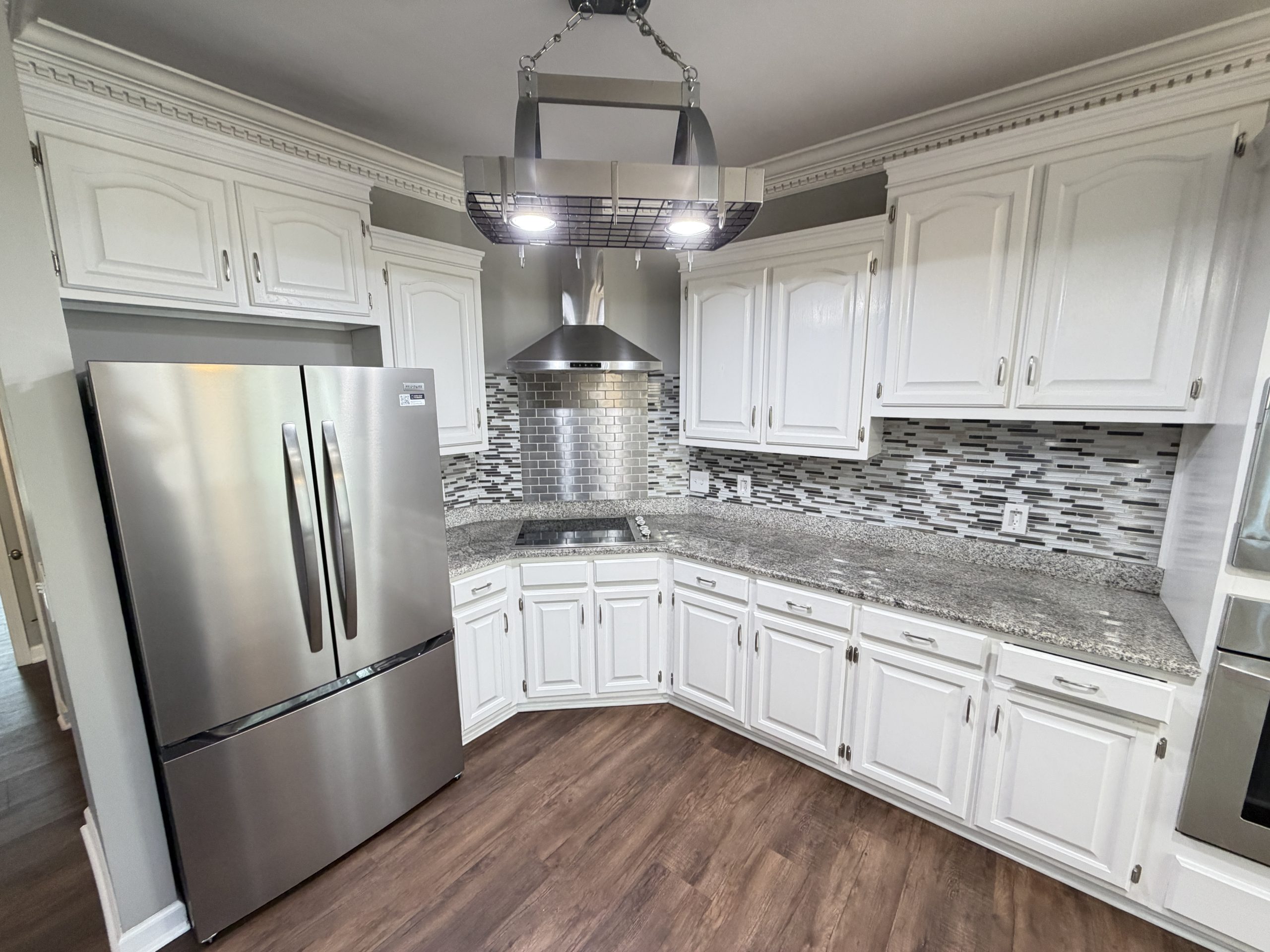 Modern kitchen with white cabinets, stainless steel refrigerator, and stainless range hood above a granite counter. A hanging pot rack with built-in lights is centered over the cooking area, with mosaic tile backsplash and dark wood flooring.