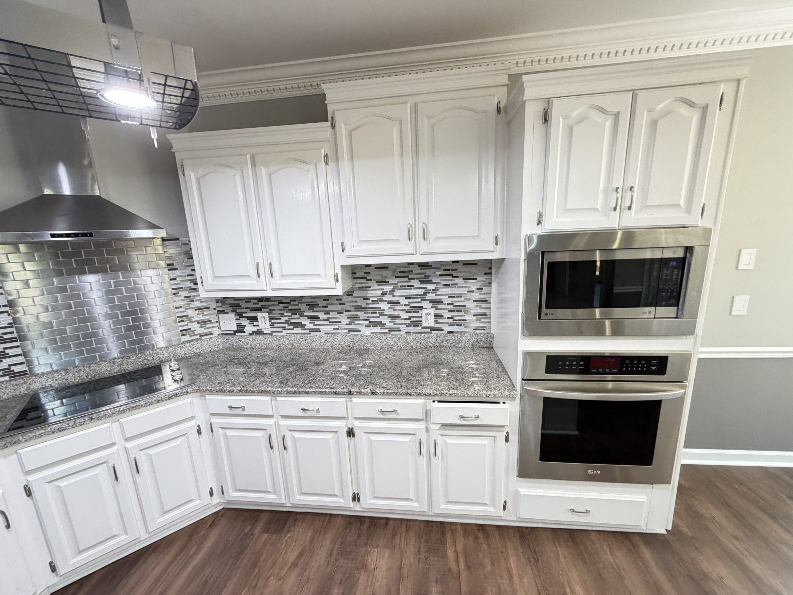 Modern kitchen with white raised-panel cabinets, stainless steel range hood, and built-in oven set against a gray-and-white mosaic tile backsplash.