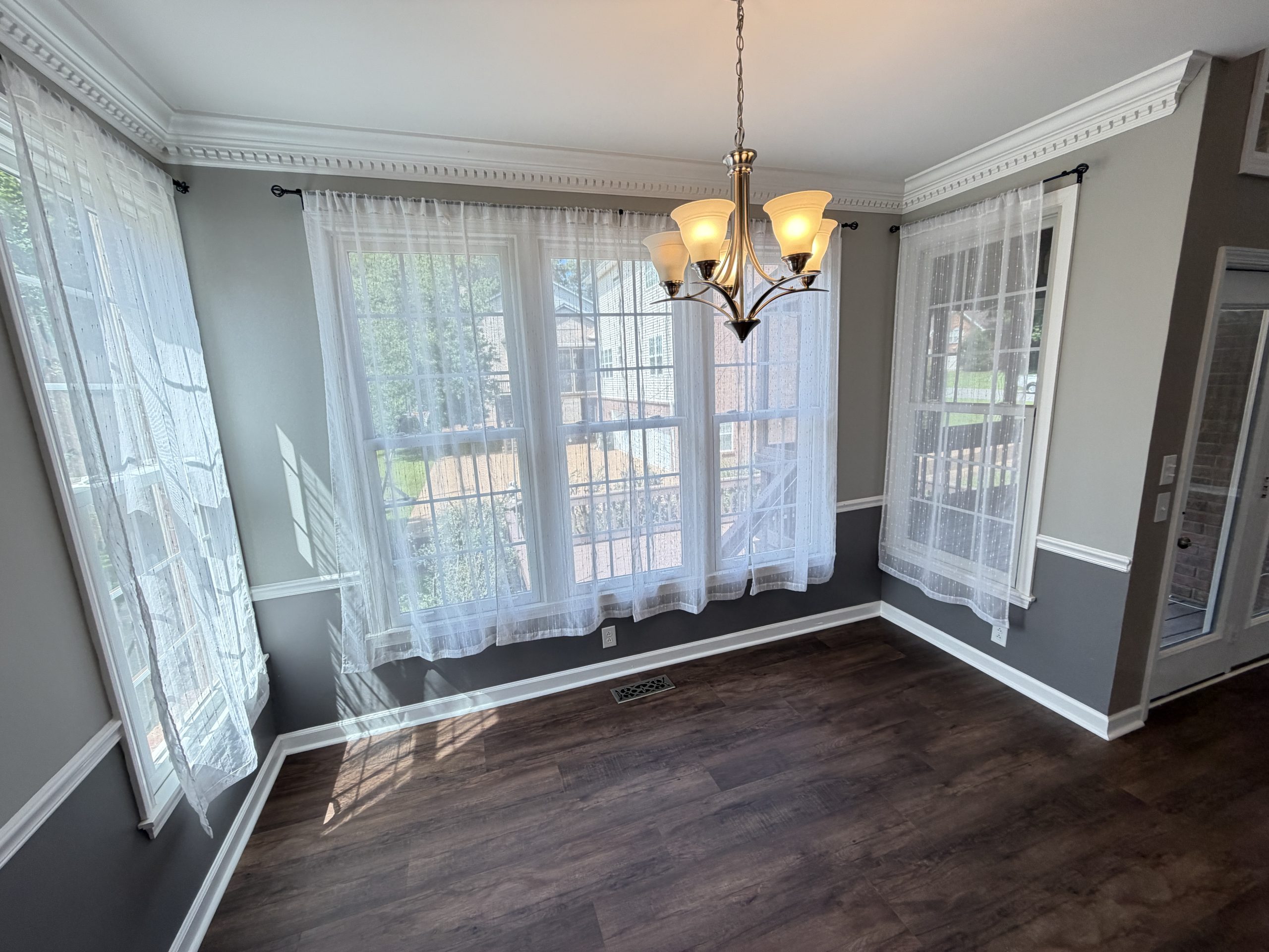 Bright dining nook with large windows and sheer white curtains, a hanging chandelier, and dark wood floor.