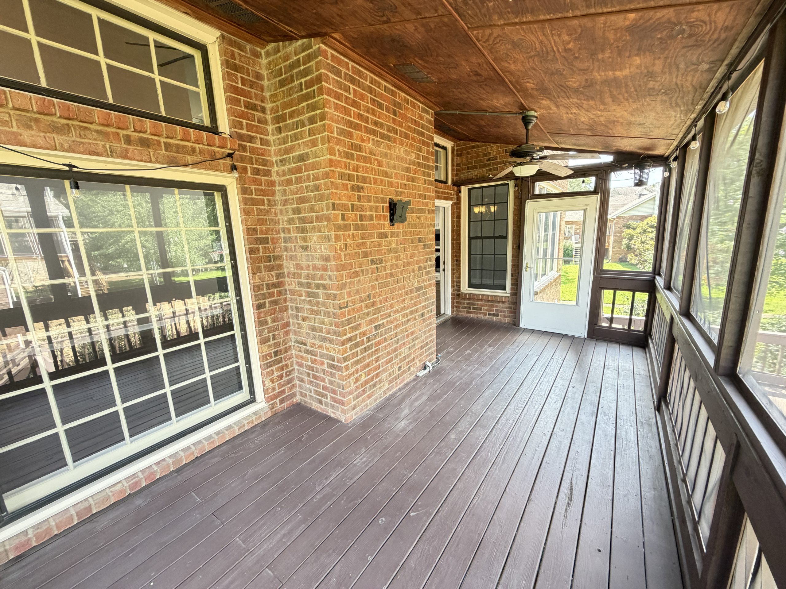 Covered brick porch with a wooden deck, string lights, and a ceiling fan, view of windows and back door.
