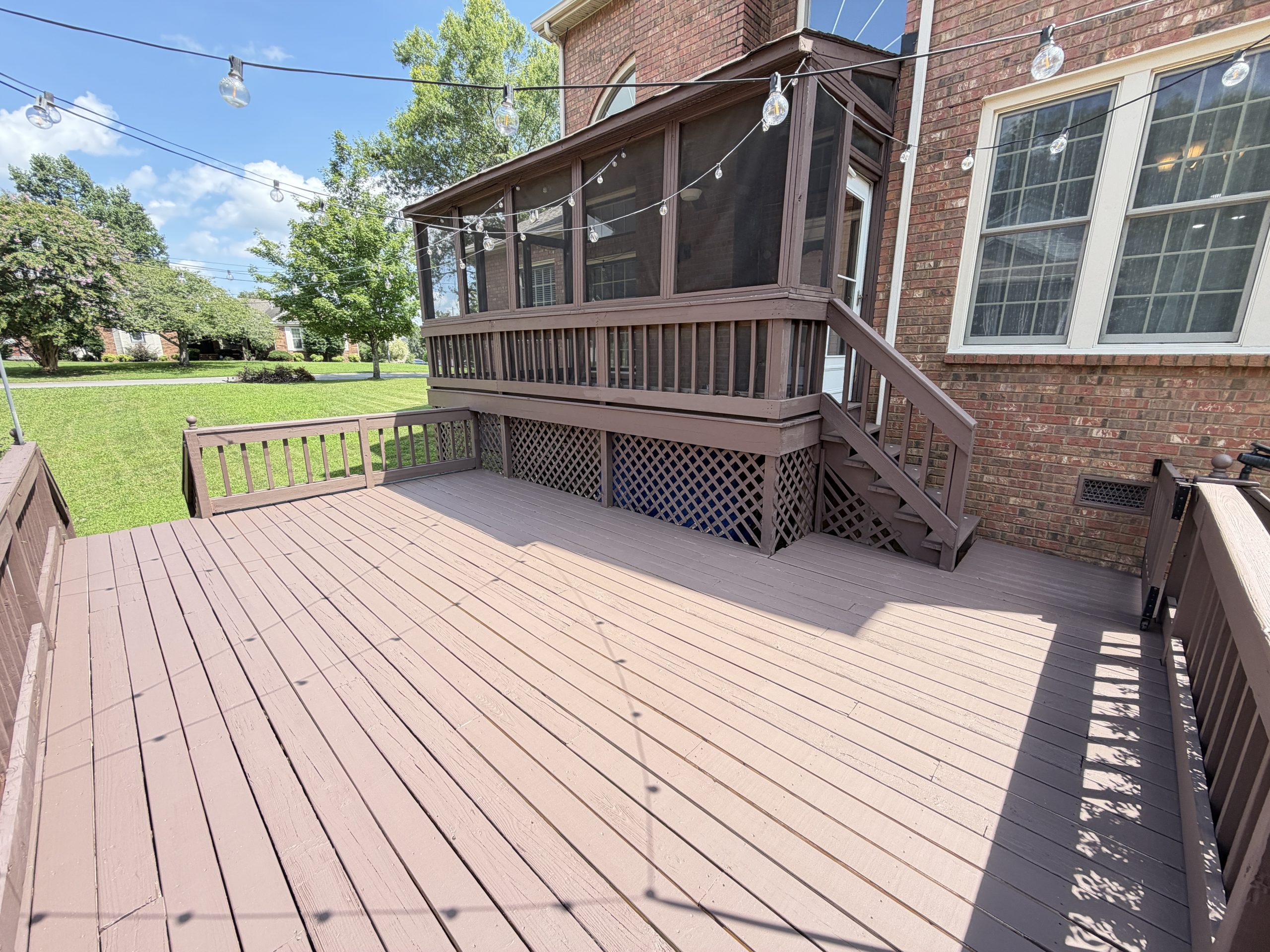 Wooden backyard deck attached to a brick house with a screened porch, string lights overhead, and a green lawn beyond.
