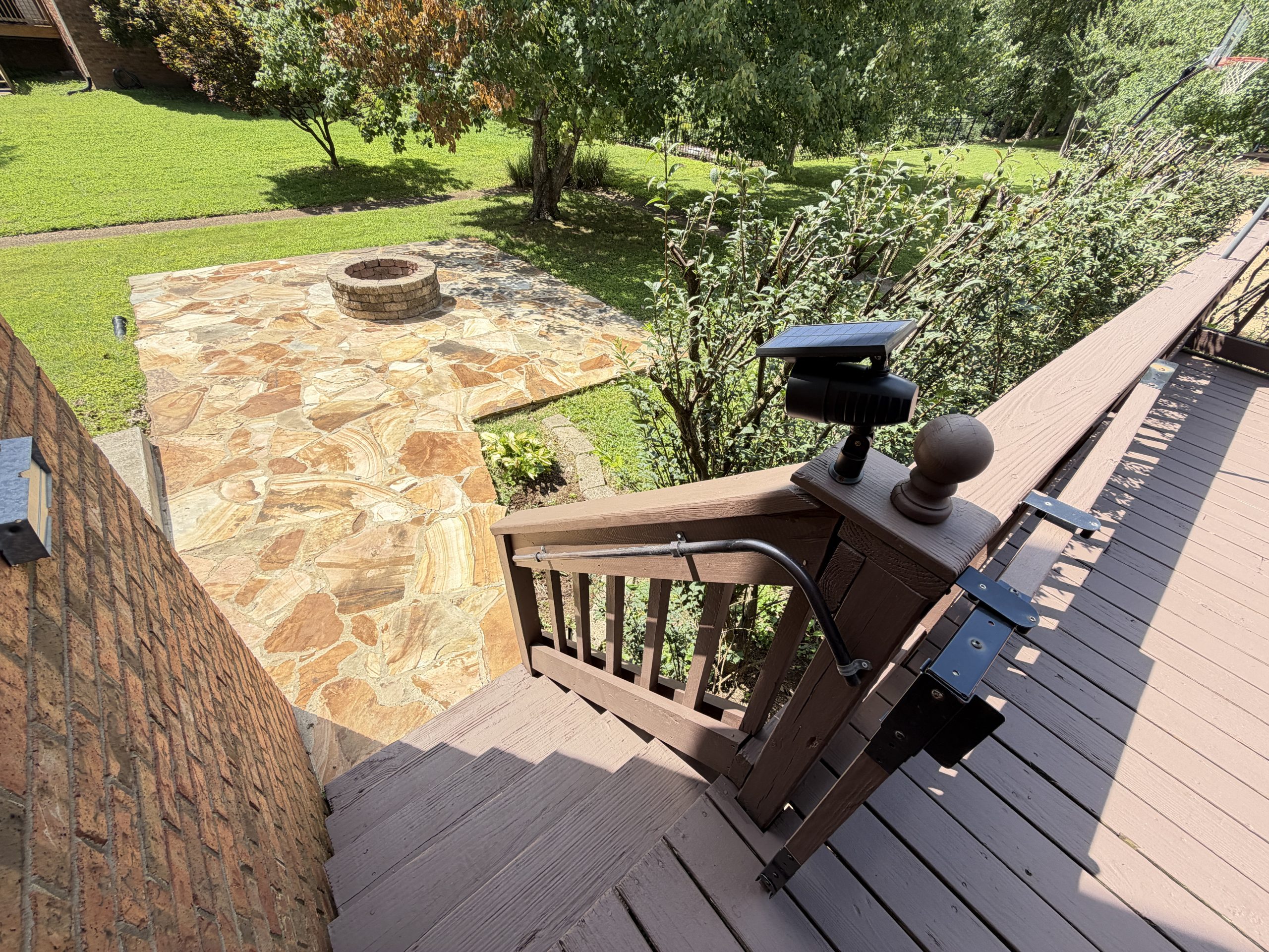 Backyard scene showing a stone patio with a circular brick fire pit, a green lawn, and trees, viewed from a wooden deck railing with outdoor equipment in the foreground.