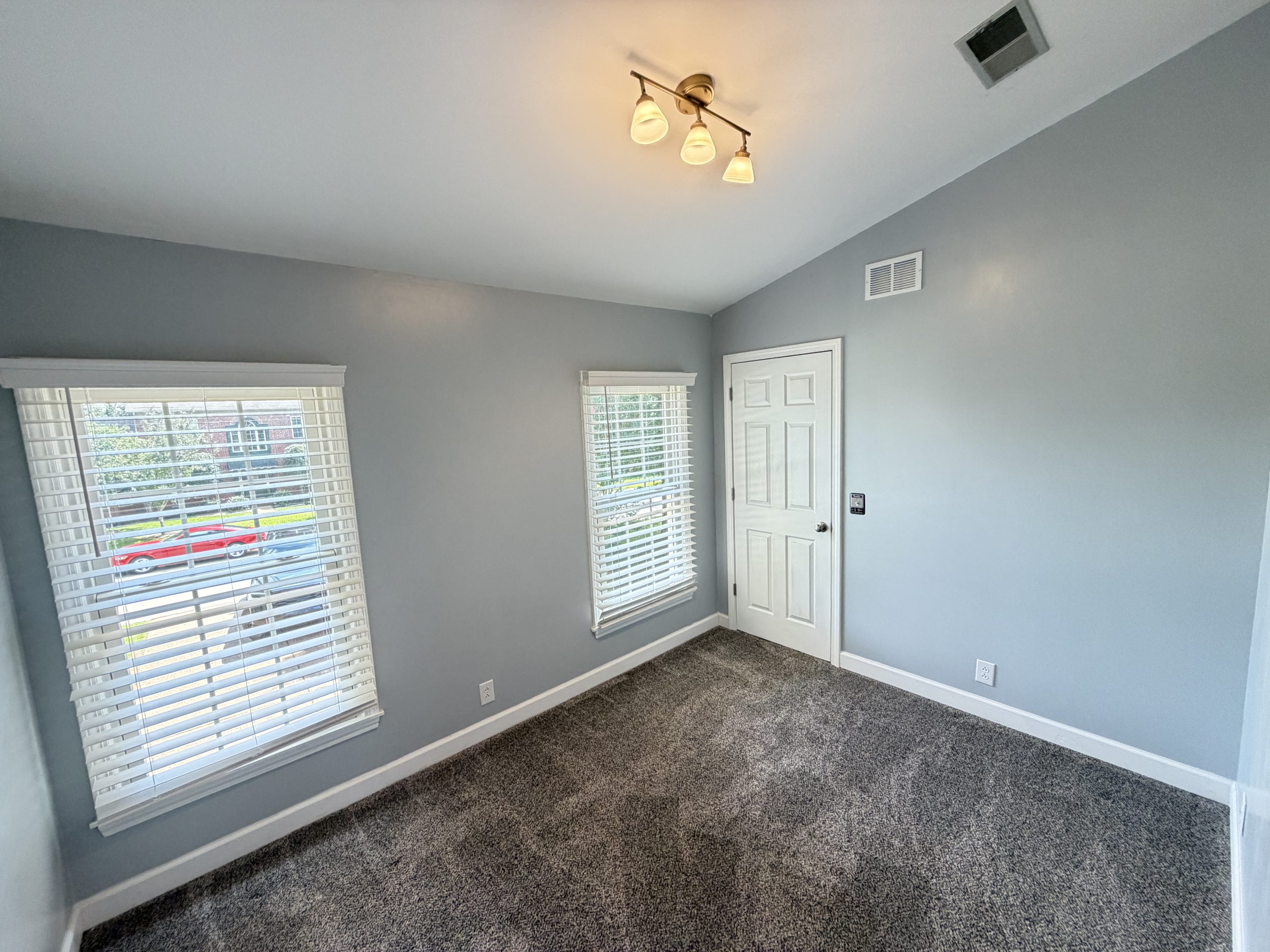 Empty light-blue room with two windows and blinds, white door, and gray carpeted floor; ceiling light fixture visible on the ceiling.