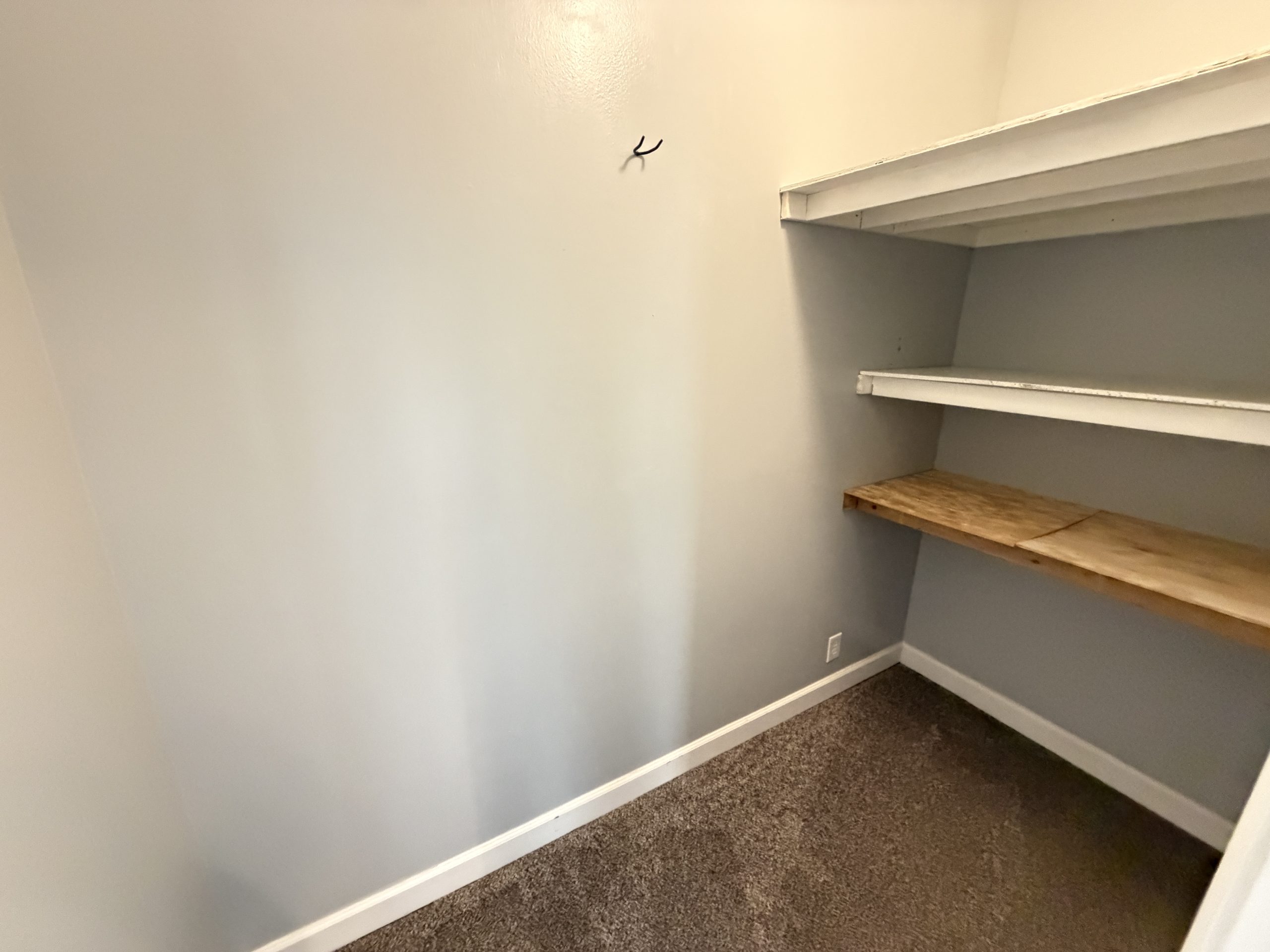 Narrow storage closet with gray walls and white built-in shelves on the right; a wooden shelf at the bottom. A small wall hook is visible.