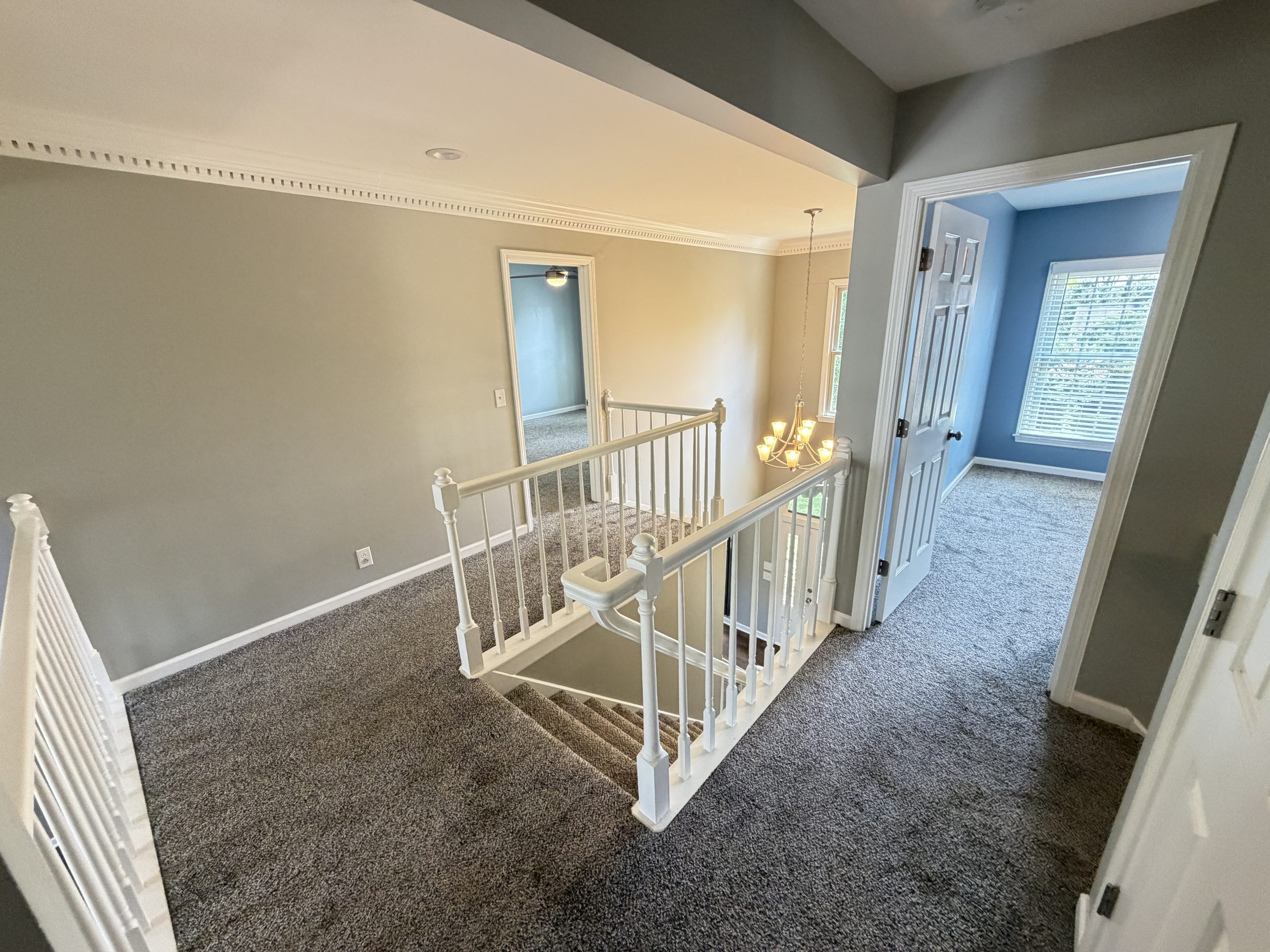 Upper-floor hallway with beige walls, white railing, and a hanging chandelier overlooking carpeted stairs toward a blue room with a window.