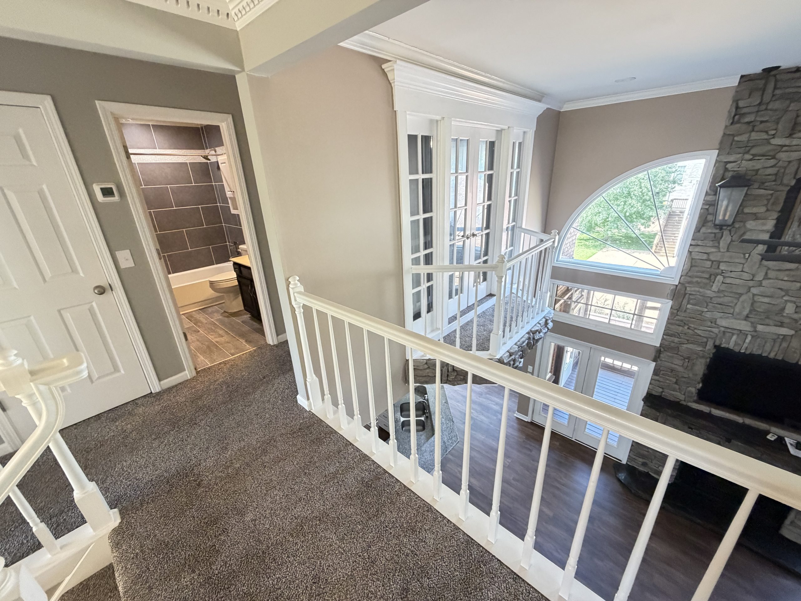 Interior of a multi-level home: carpeted hallway with white railings, stone fireplace to the right, and a large arched window with grid panes letting in light.