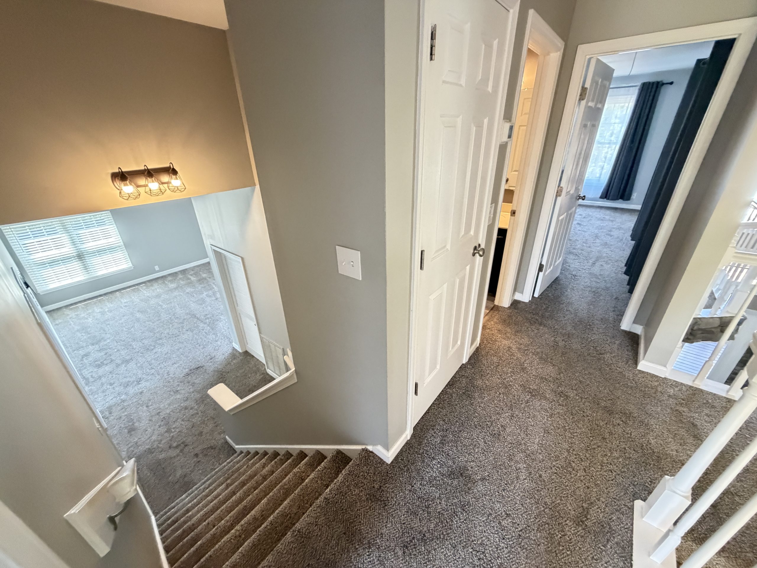 View down a carpeted staircase from the upper landing into a hallway with beige walls and white trim, several closed doors visible along a narrow corridor.