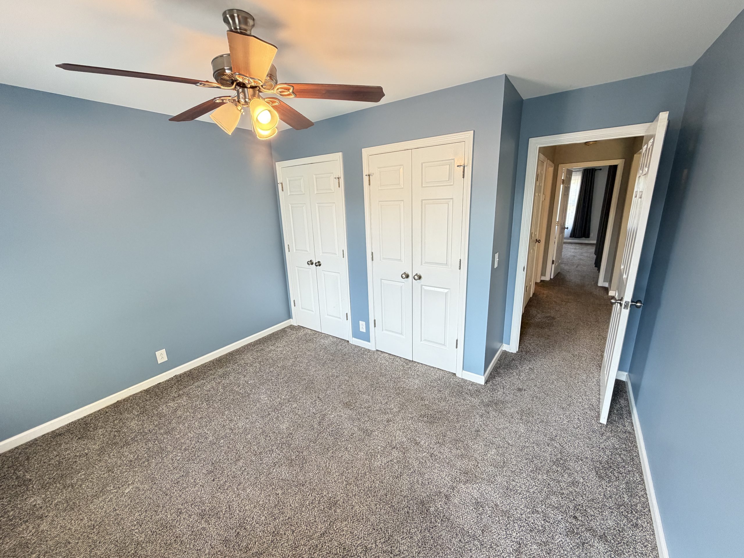 Blue bedroom with white six-panel closet doors, beige carpet, and a ceiling fan overhead.
