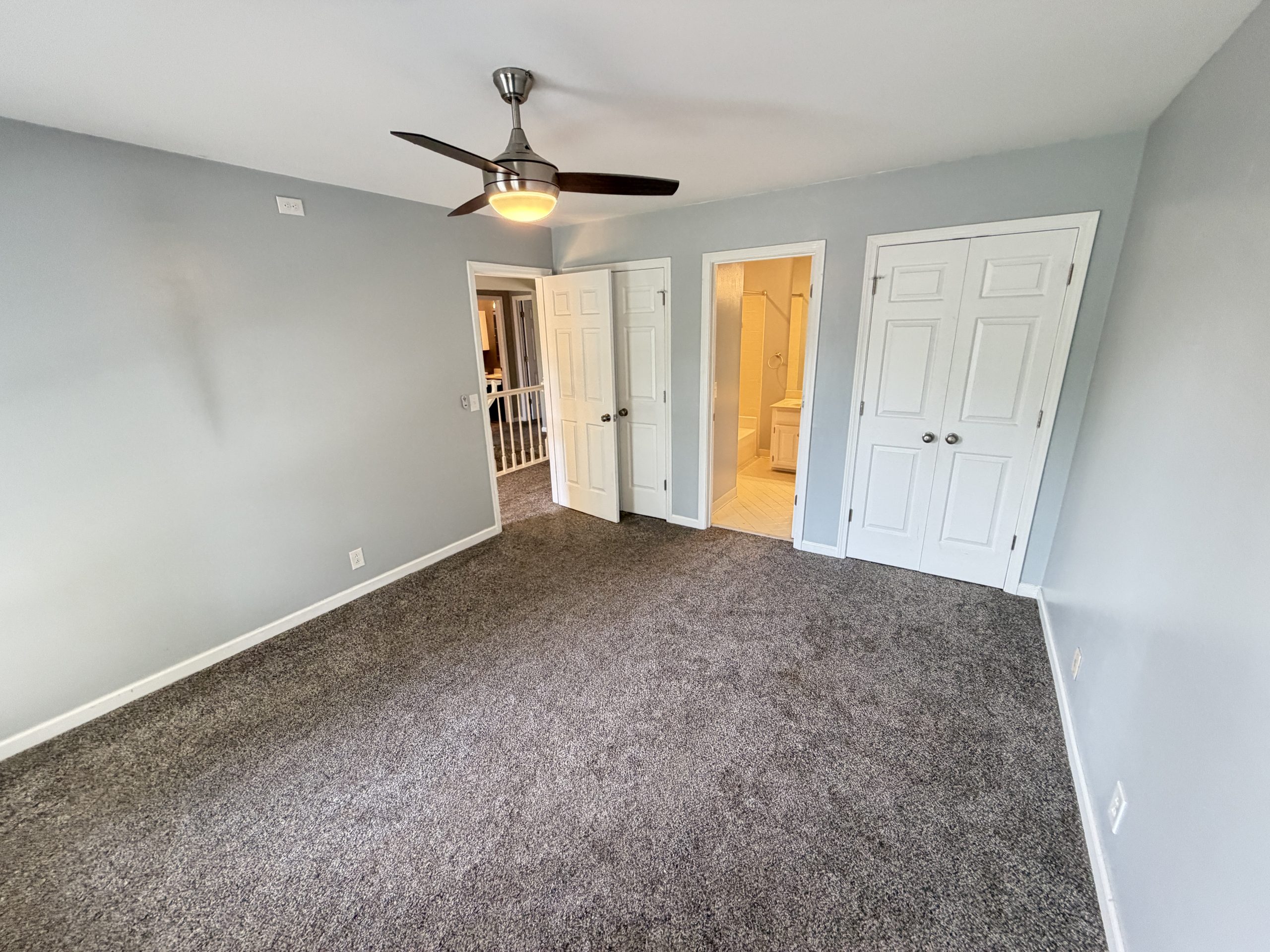 Empty bedroom with gray walls, brown carpet, and a modern ceiling fan; multiple white doors line the wall, one open to a bathroom area