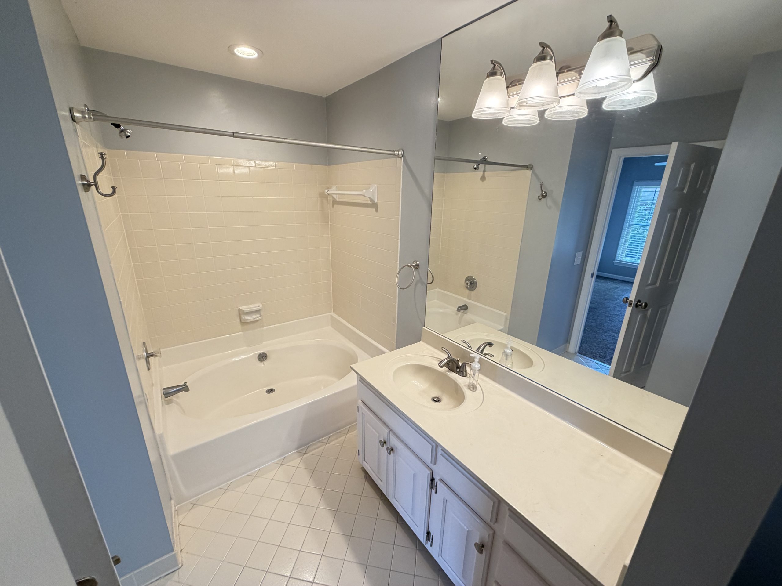 Blue bathroom with a white tile shower/tub, a long white double-sink vanity, and a large mirror with four-light fixture above.