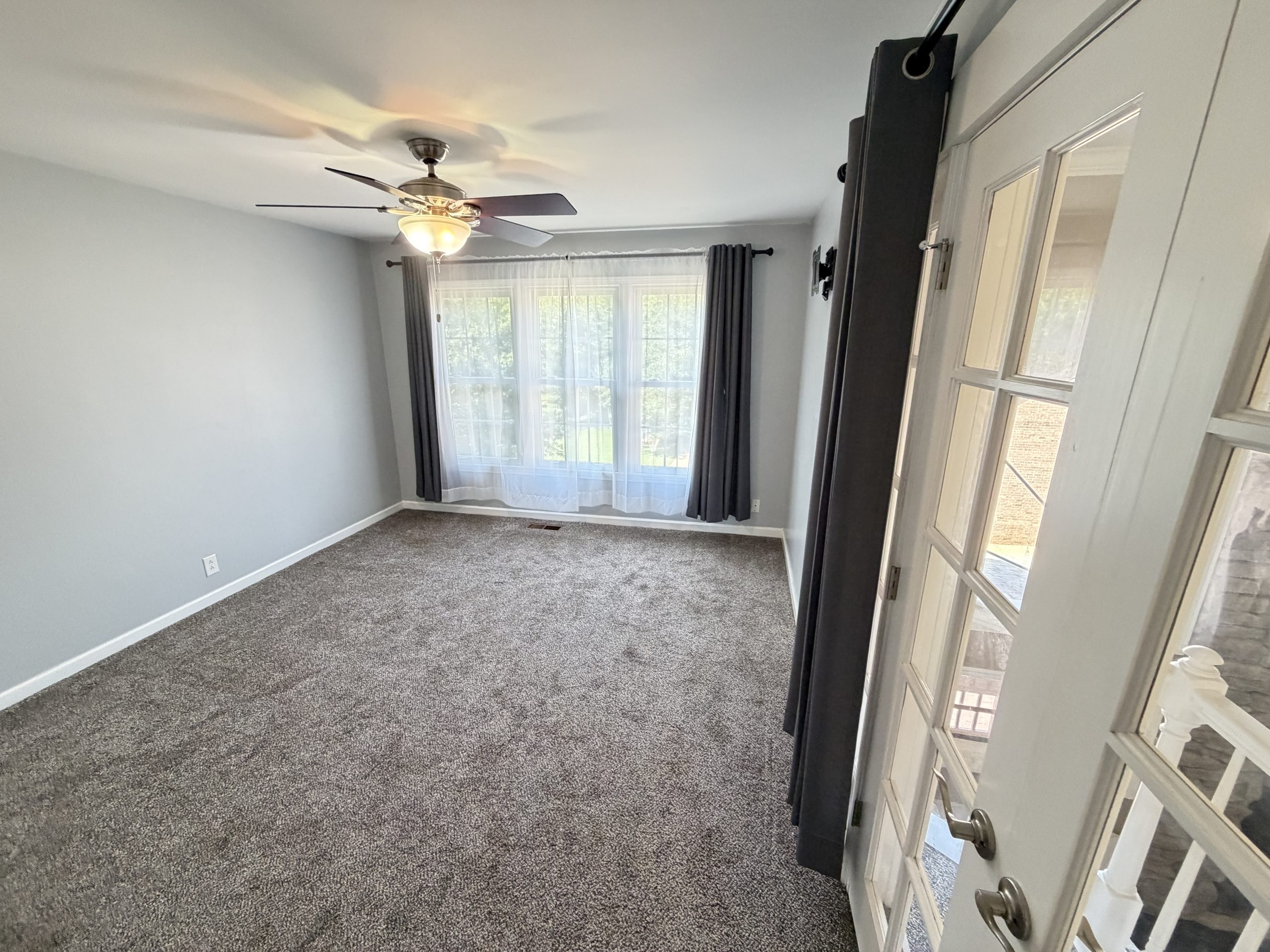 Empty gray bedroom with a ceiling fan and light, large window with sheer curtains and dark drapes, door with glass panels nearby