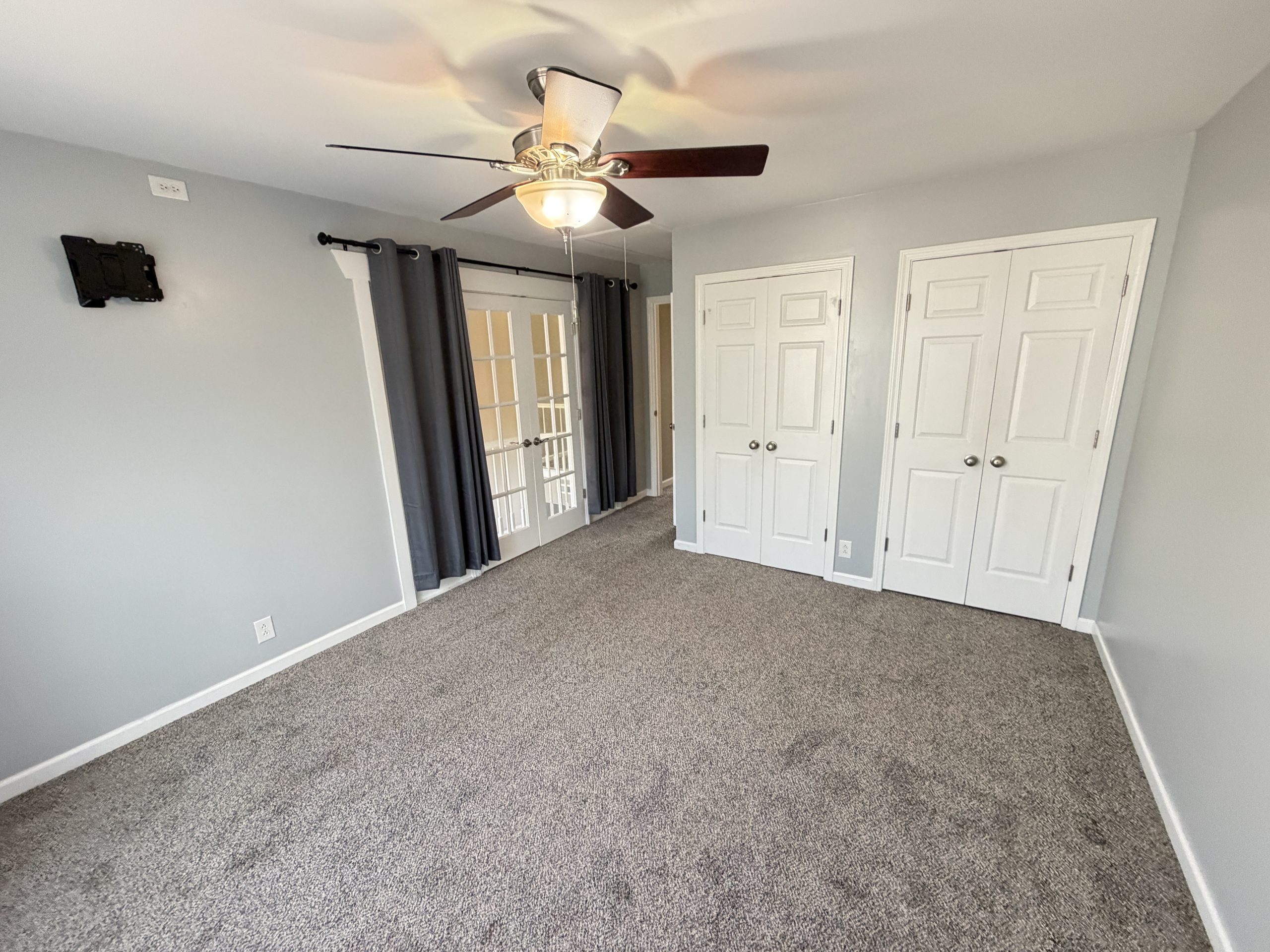 Empty living room with gray walls, a ceiling fan, and white double doors; dark curtains cover glass French doors in the background.