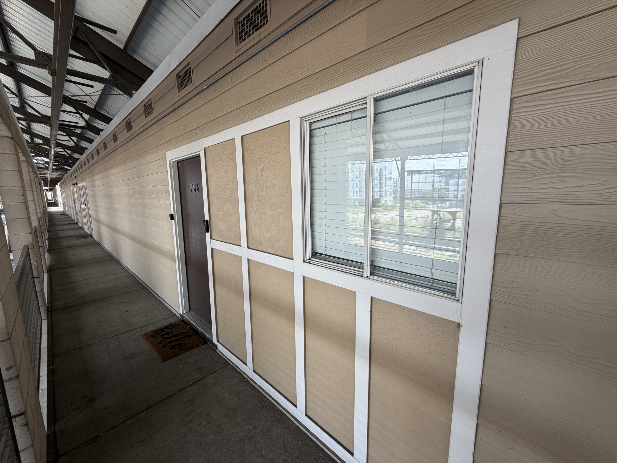 Exterior apartment corridor with beige siding, white trim, and a metal door beside a large window with blinds in a long walkway.