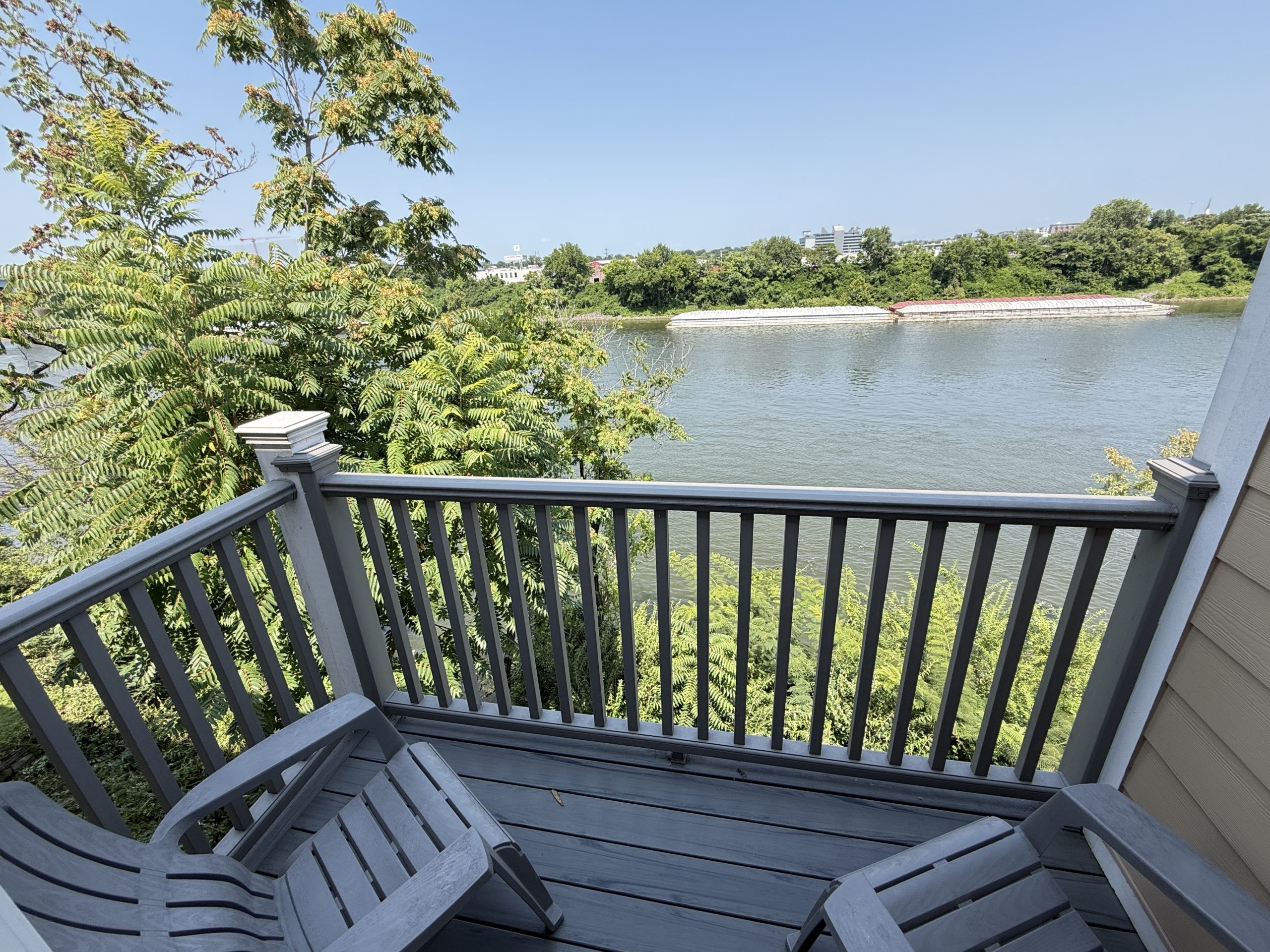 Wooden balcony with gray railing and stairs overlooking a calm river, banks lined with green trees and distant buildings on the far shore.