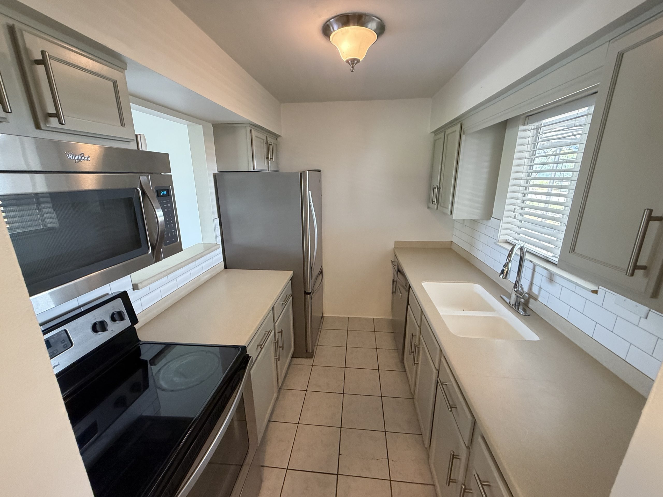 Narrow galley kitchen with white cabinets, beige countertops, and stainless steel refrigerator and oven. Window with blinds at the far end.