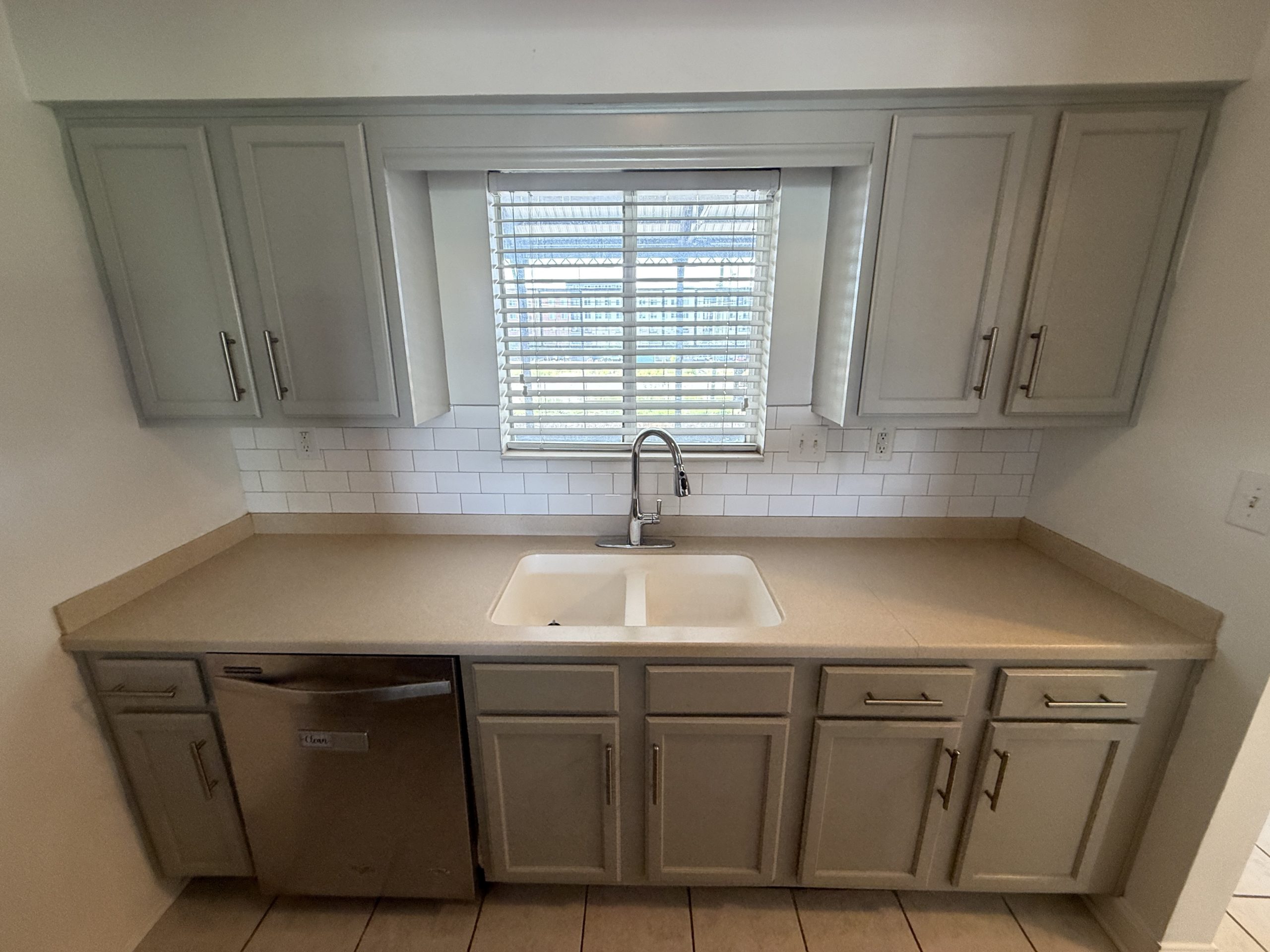 Grey upper and lower cabinets surround a window over a sink with a chrome faucet. Beneficial lighting from the window adds brightness to the kitchen workspace.