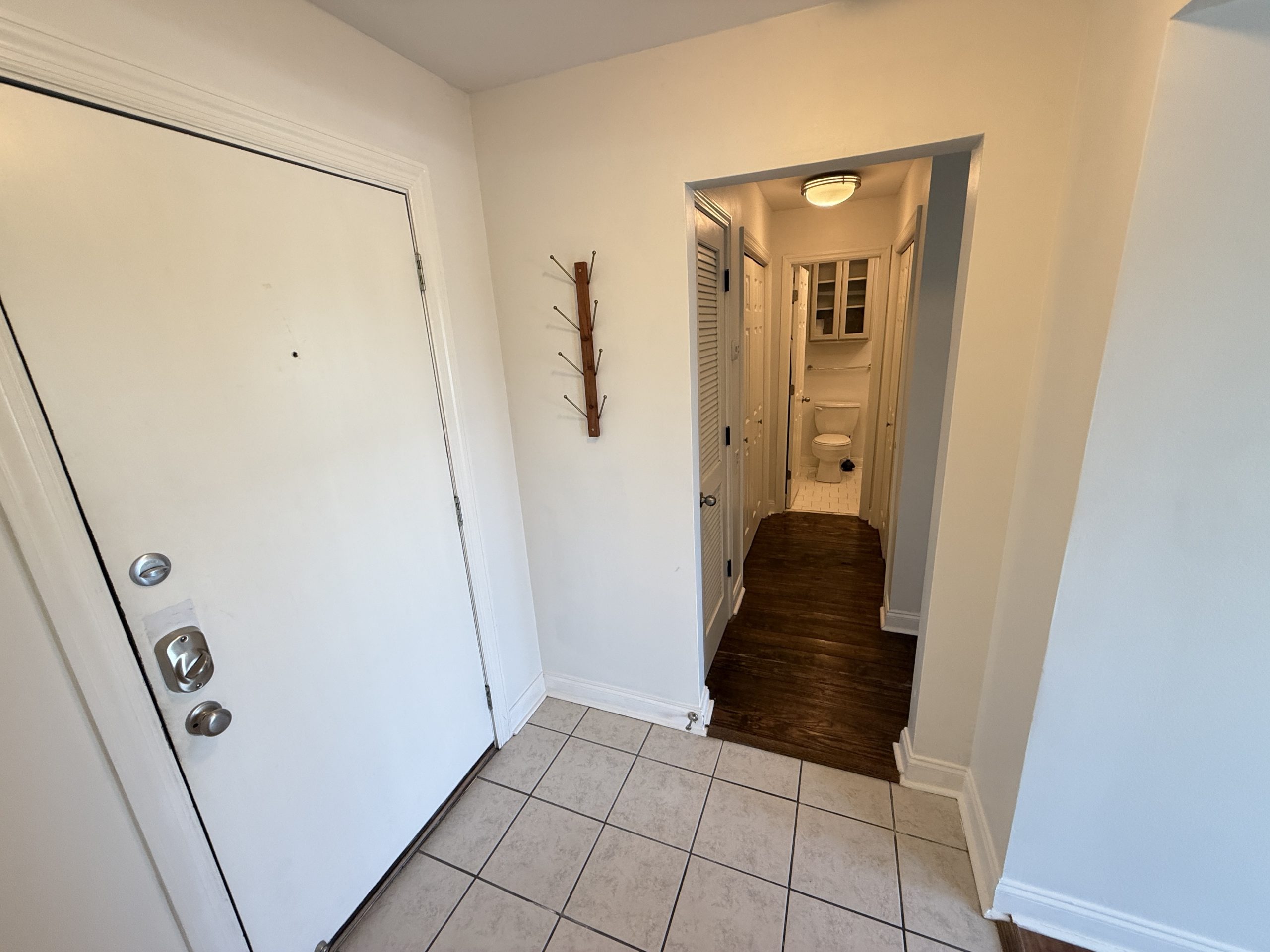 Narrow white entry hallway with a tiled floor, a wooden coat rack on the wall, and a view down to a bathroom at the end.