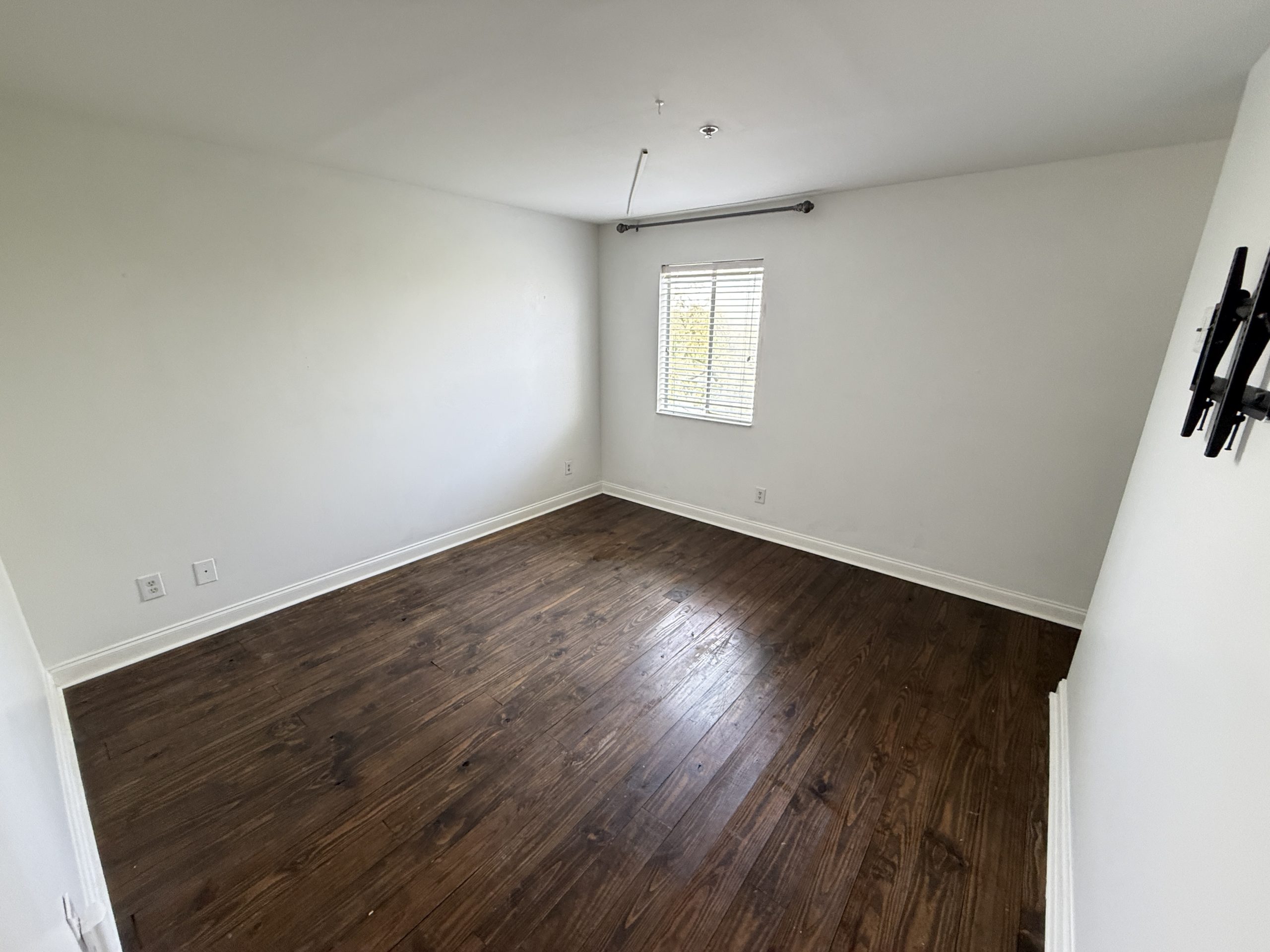 Empty room with dark wooden floor, white walls, a small window with blinds, and a wall-mounted TV bracket on the right side.