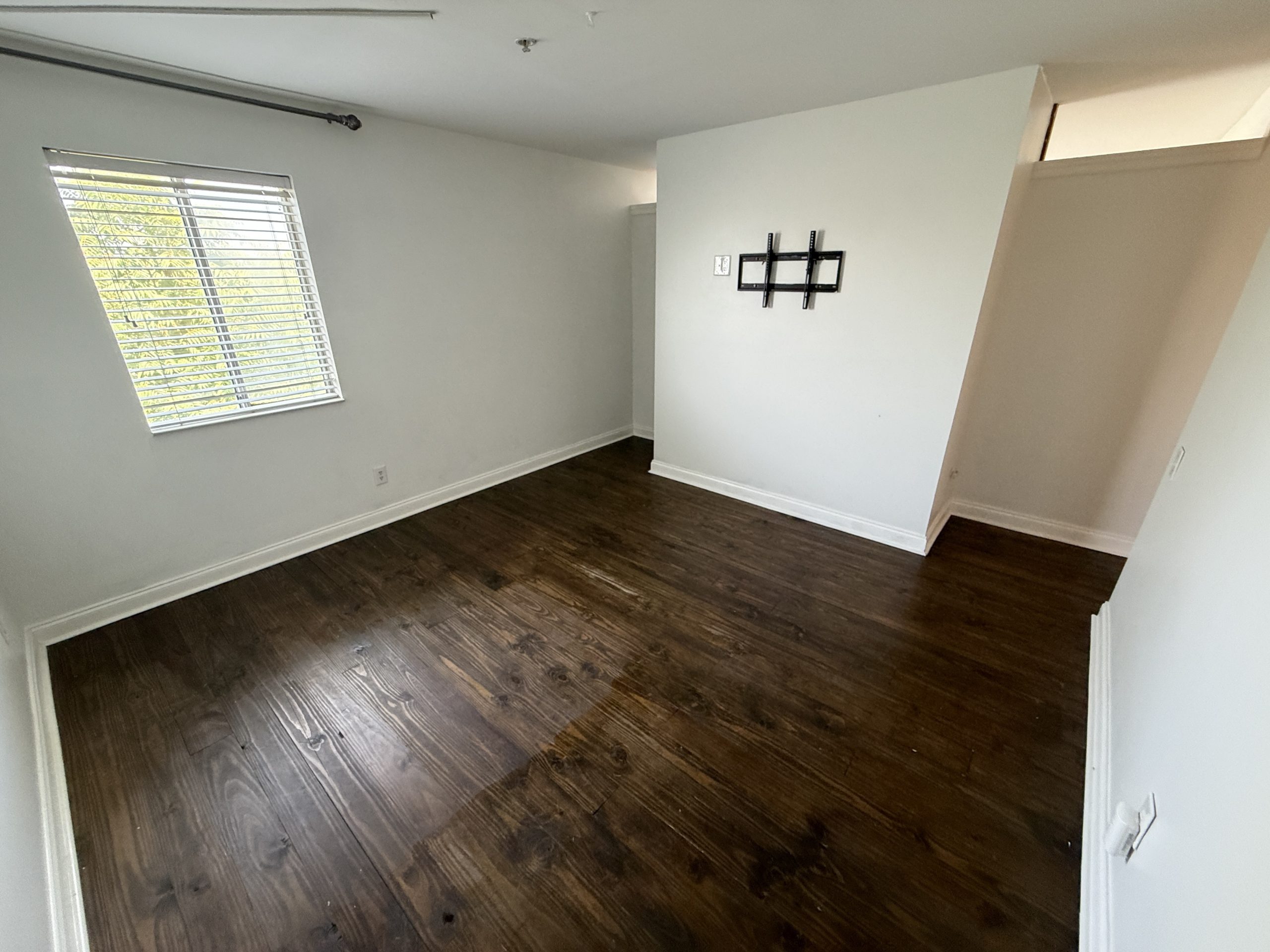 Empty room with dark wood floors, white walls, and a window with horizontal blinds on the left side.