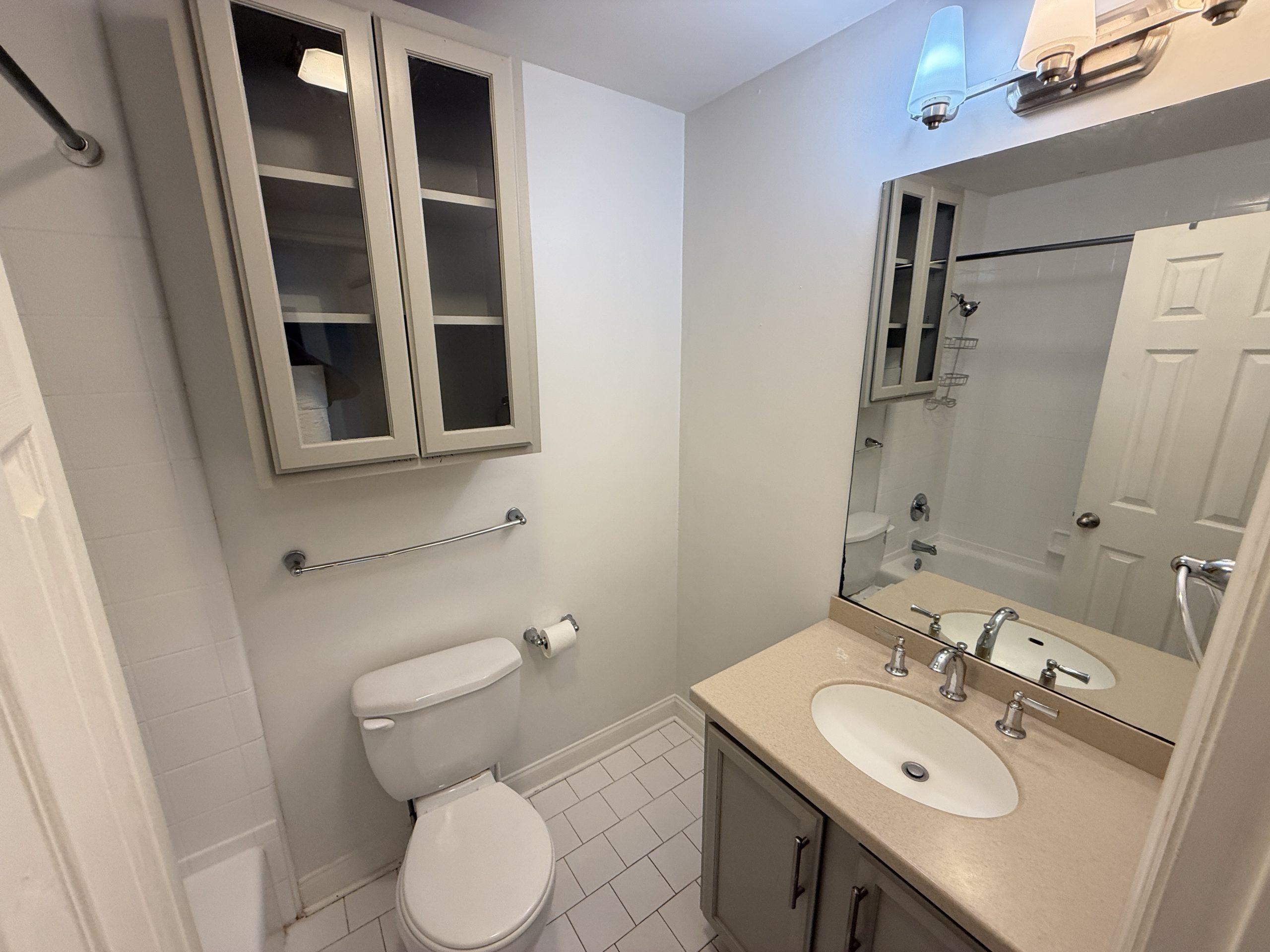 Small bathroom with a double-sink vanity, beige countertop, and a large wall mirror reflecting a tub/shower area.