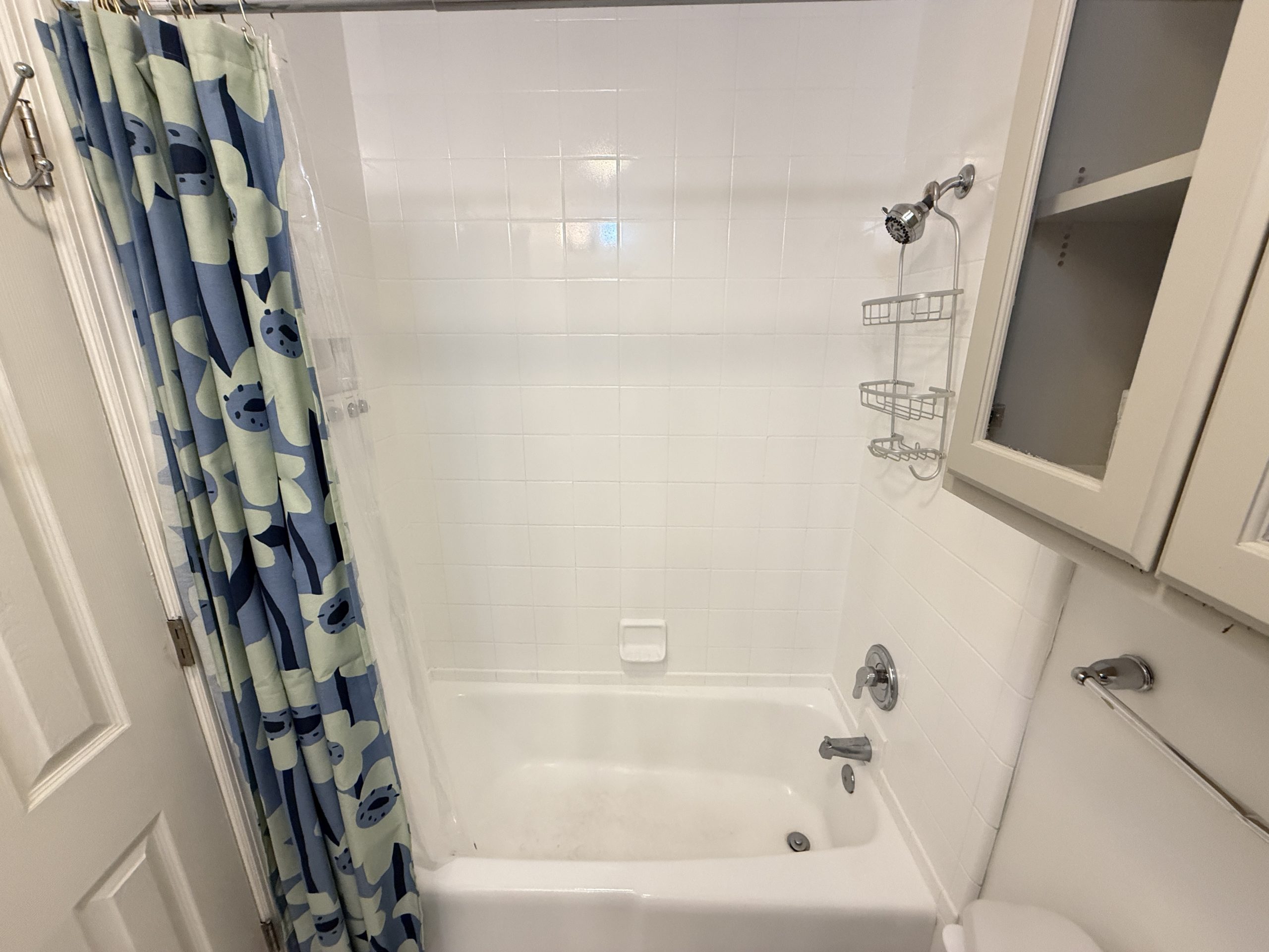 Bathroom shower with blue patterned curtain, white tiled walls, and a hanging metal shower rack over a tub/shower combo