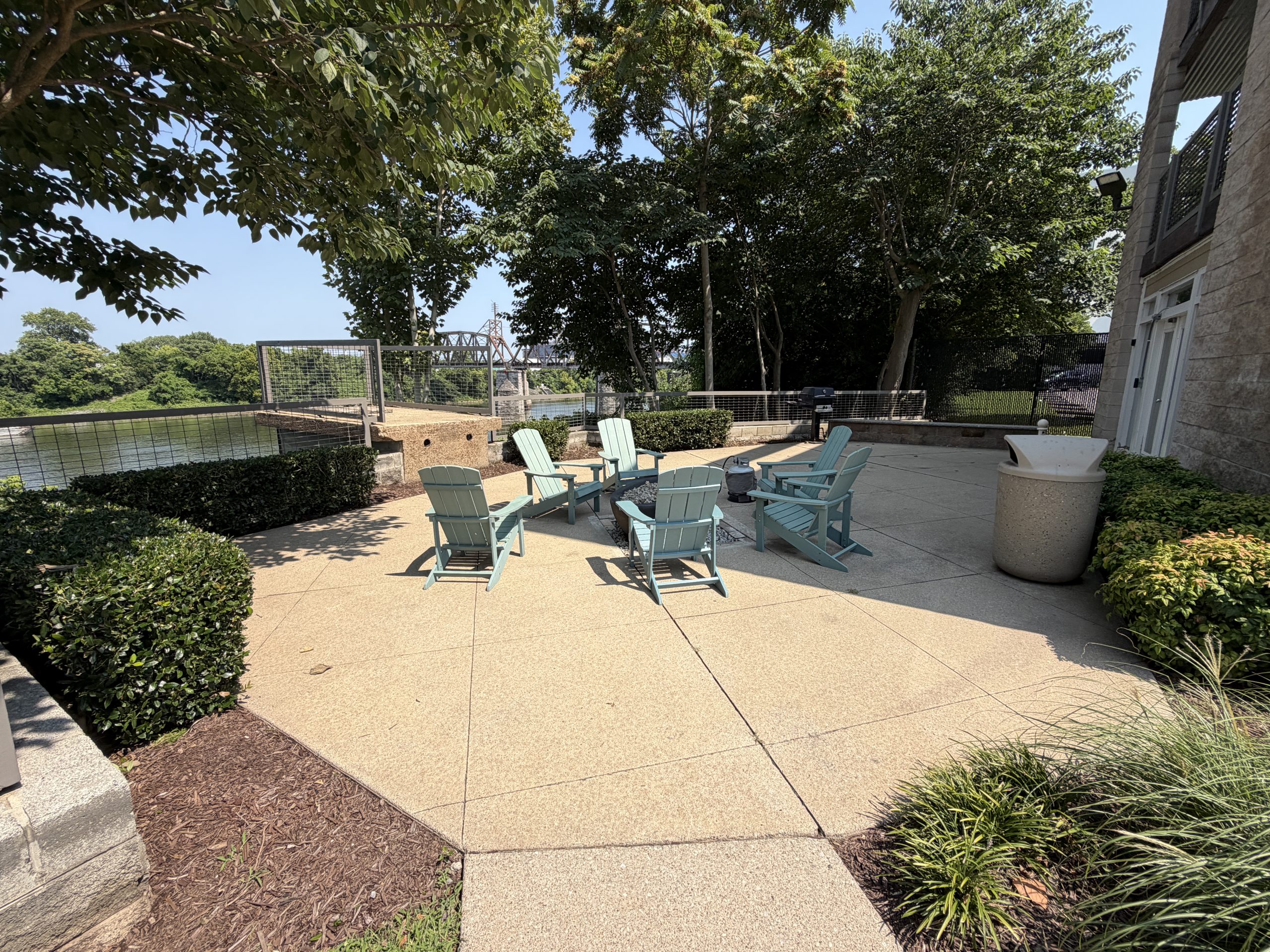 Outdoor patio with light blue Adirondack chairs arranged around a fire pit by a lake, surrounded by trees and a stone building on the right side.