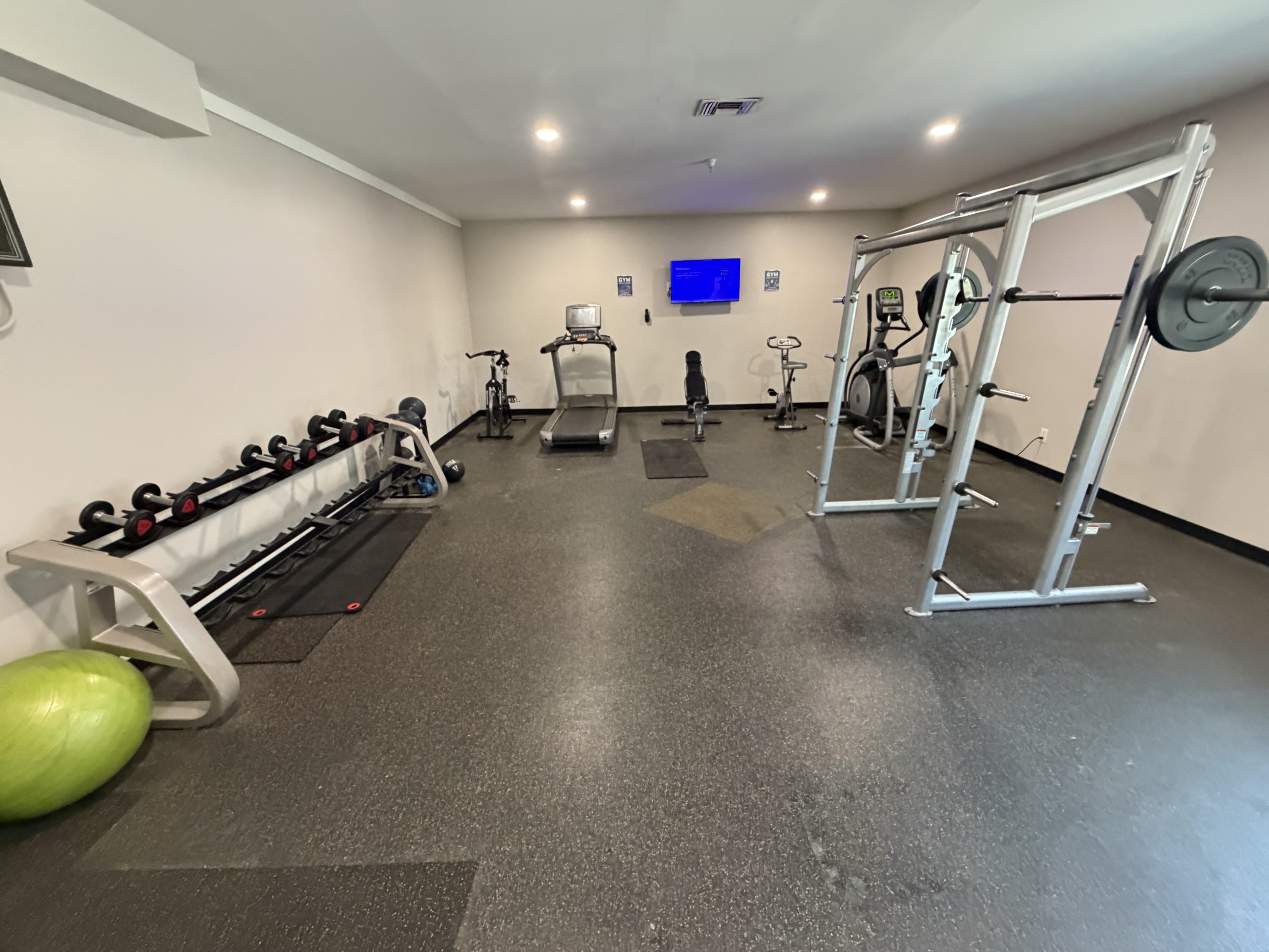 Gym interior with free weights on a rack along the left wall and a large lime-green stability ball in the foreground; cardio machines and a squat rack are visible on the right.