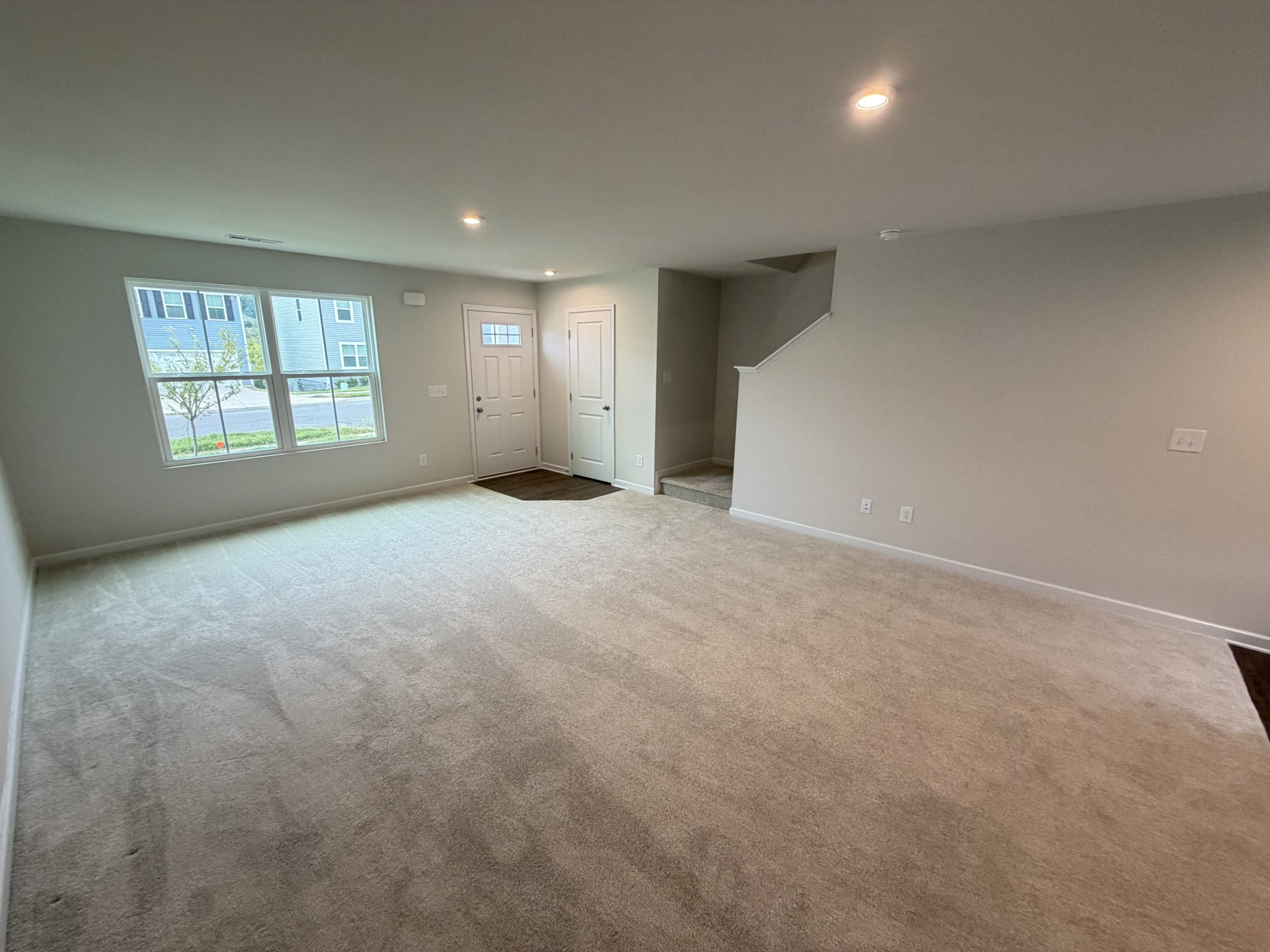 Empty living room with beige carpet, gray walls, a large window, front door, and stairway to the right.