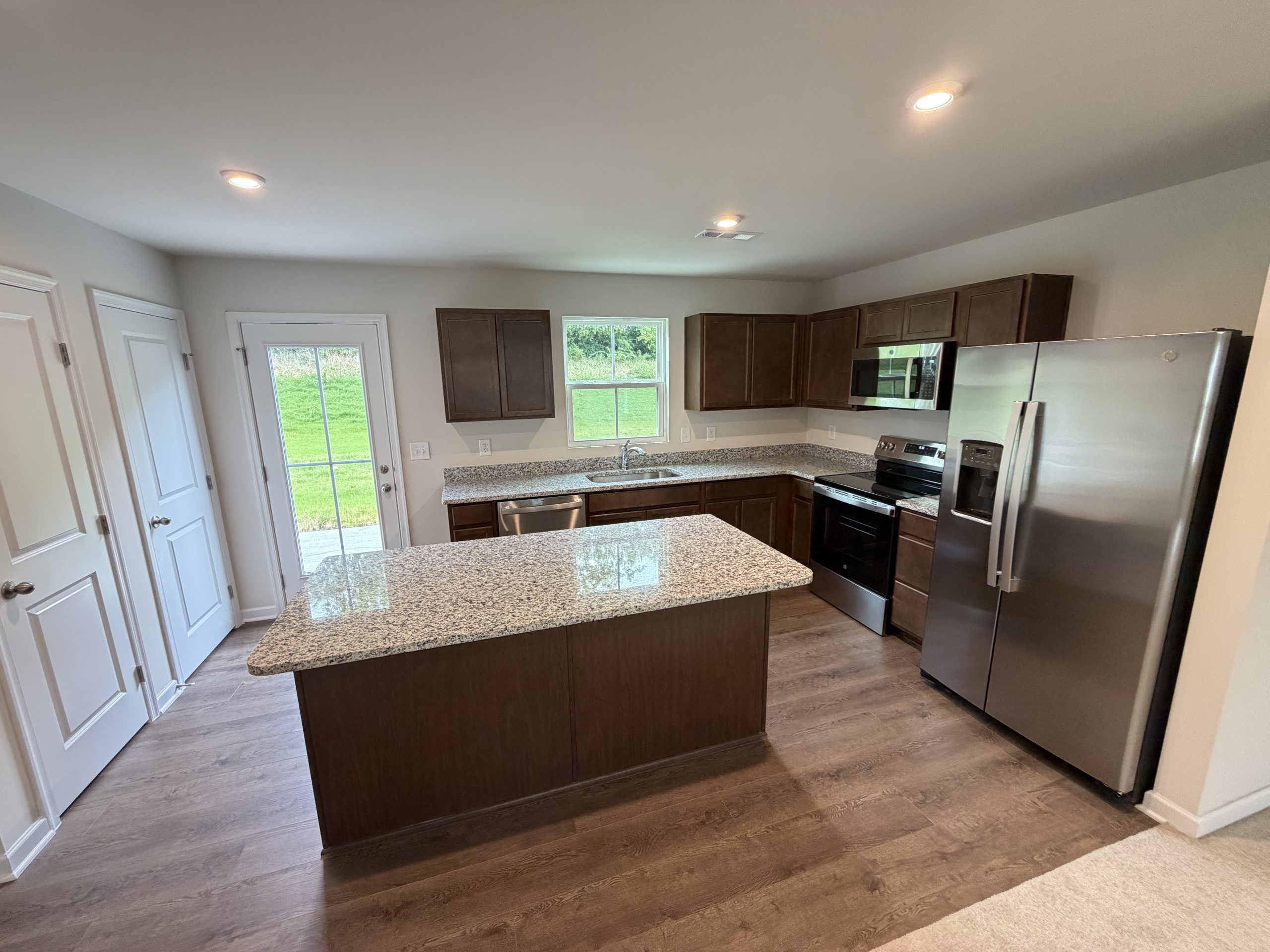 Modern kitchen with dark wood cabinets, granite countertops, stainless steel appliances, and an island. Outside door visible to left; window above sink offers natural light.