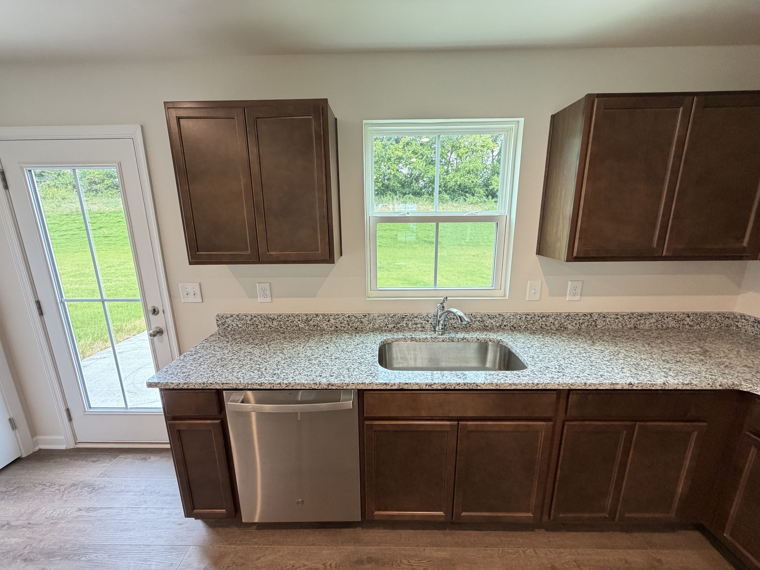 Granite kitchen counter with stainless sink and dishwasher, dark wood cabinets, window above sink, outside greenery visible.